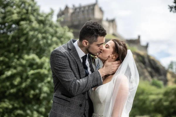 A bride and groom kissing outdoors with a Edinburgh Castle on a hill in the background - at The Caledonian Edinburgh wedding.