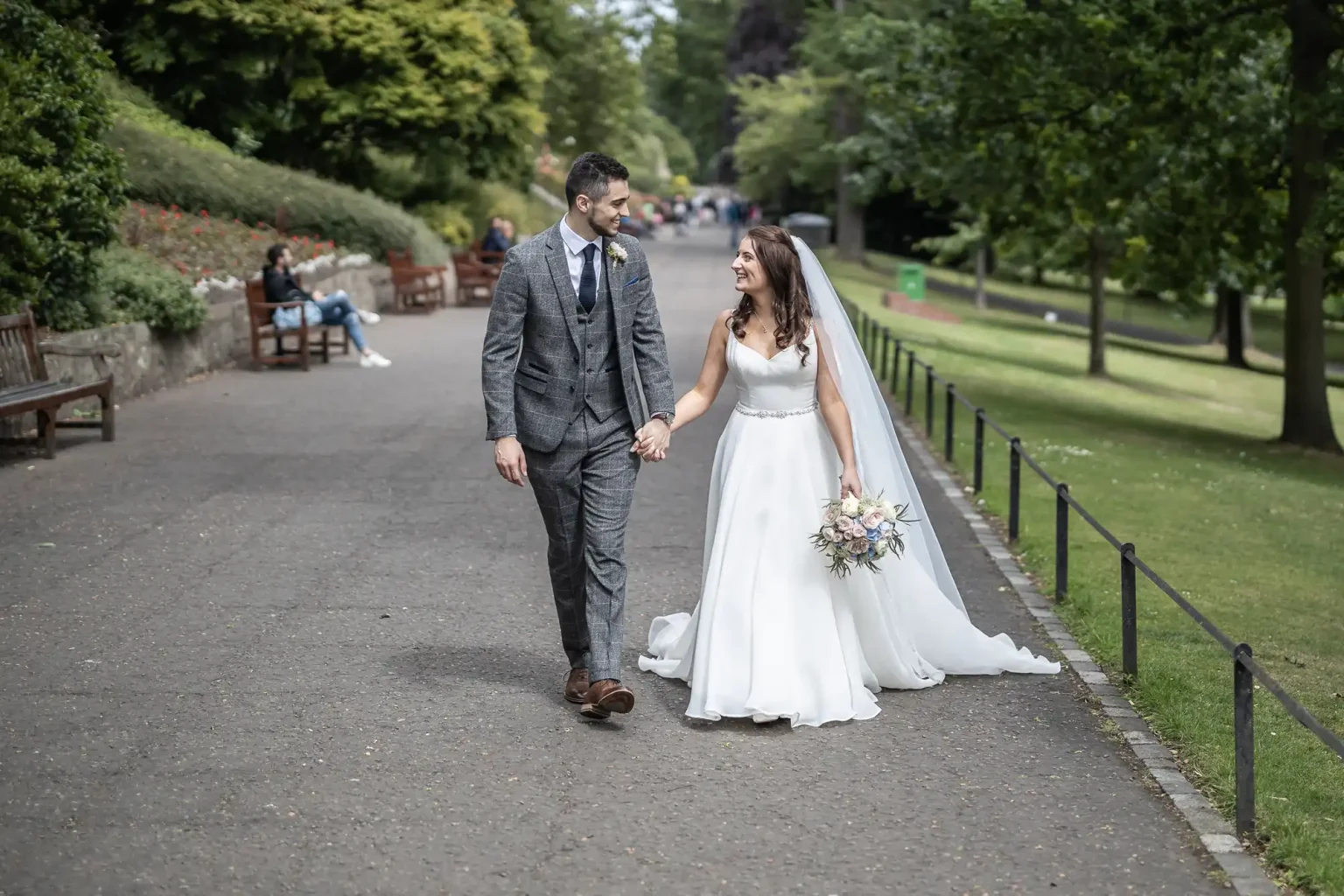 A newlywed couple walks hand in hand down a park path, smiling at each other, with the bride in a white dress and the groom in a gray suit.