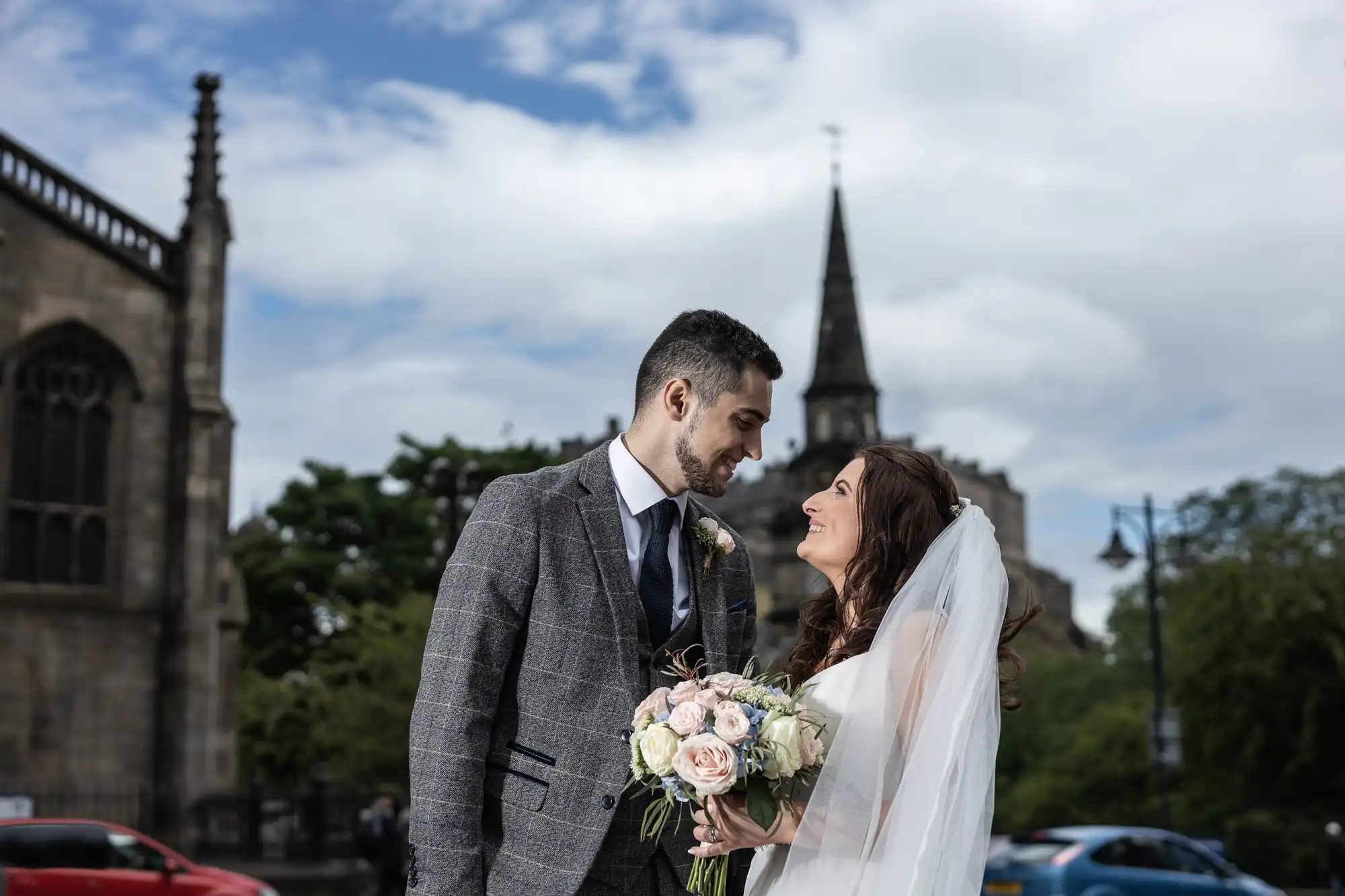 A newlywed couple gazes lovingly at each other outside, holding a bouquet, with a church and cloudy sky in the background.