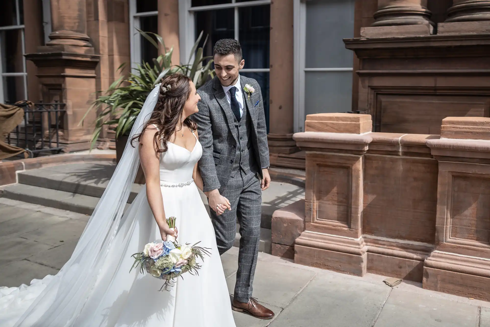 A bride in a white dress and a groom in a gray suit smiling at each other while walking next to a sandstone building.
