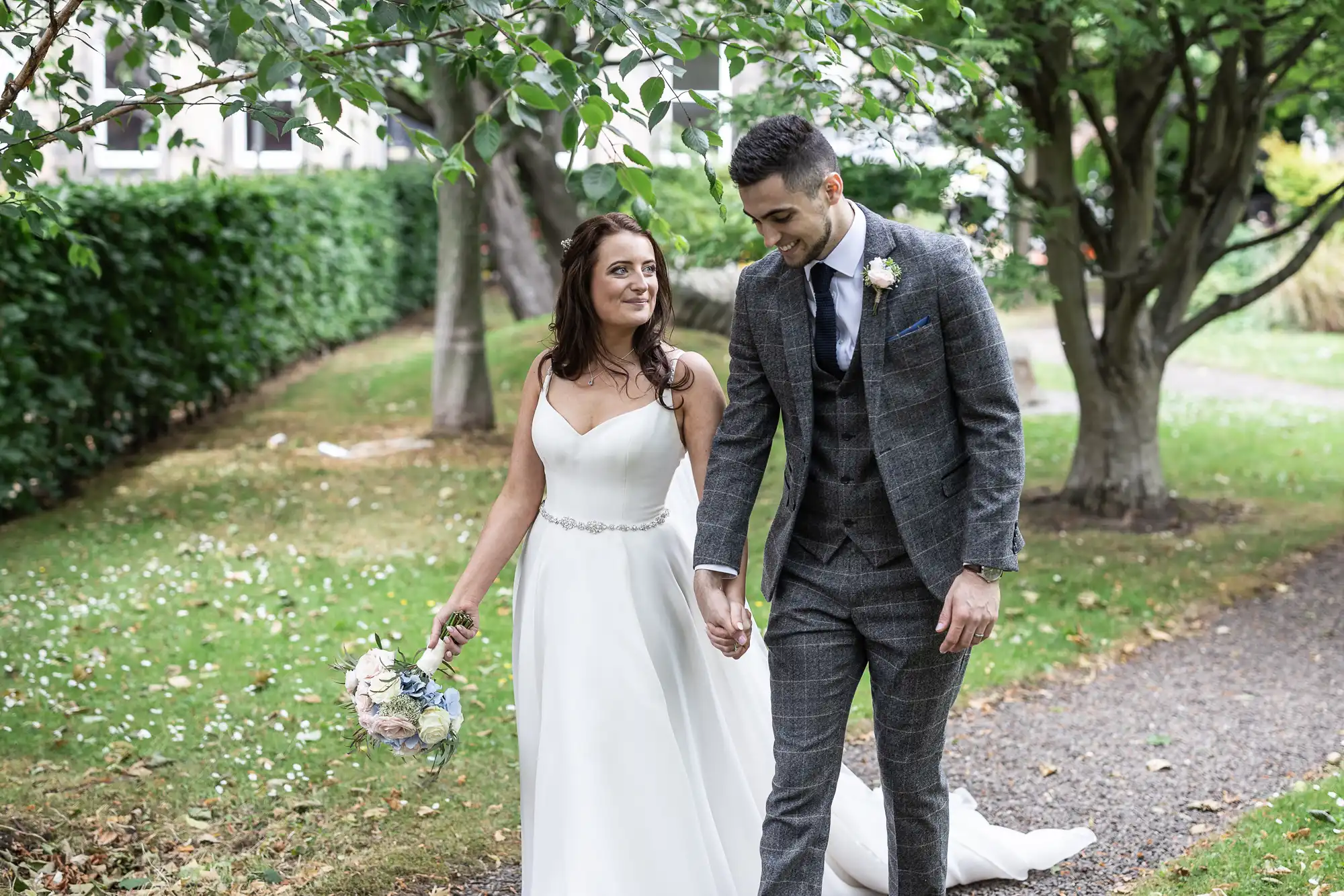A bride and groom walking hand in hand through a garden pathway, smiling at each other. The bride is holding a bouquet.