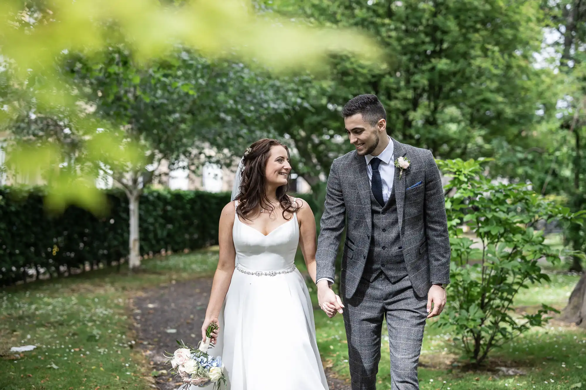 Bride and groom holding hands and smiling while walking through a lush garden.