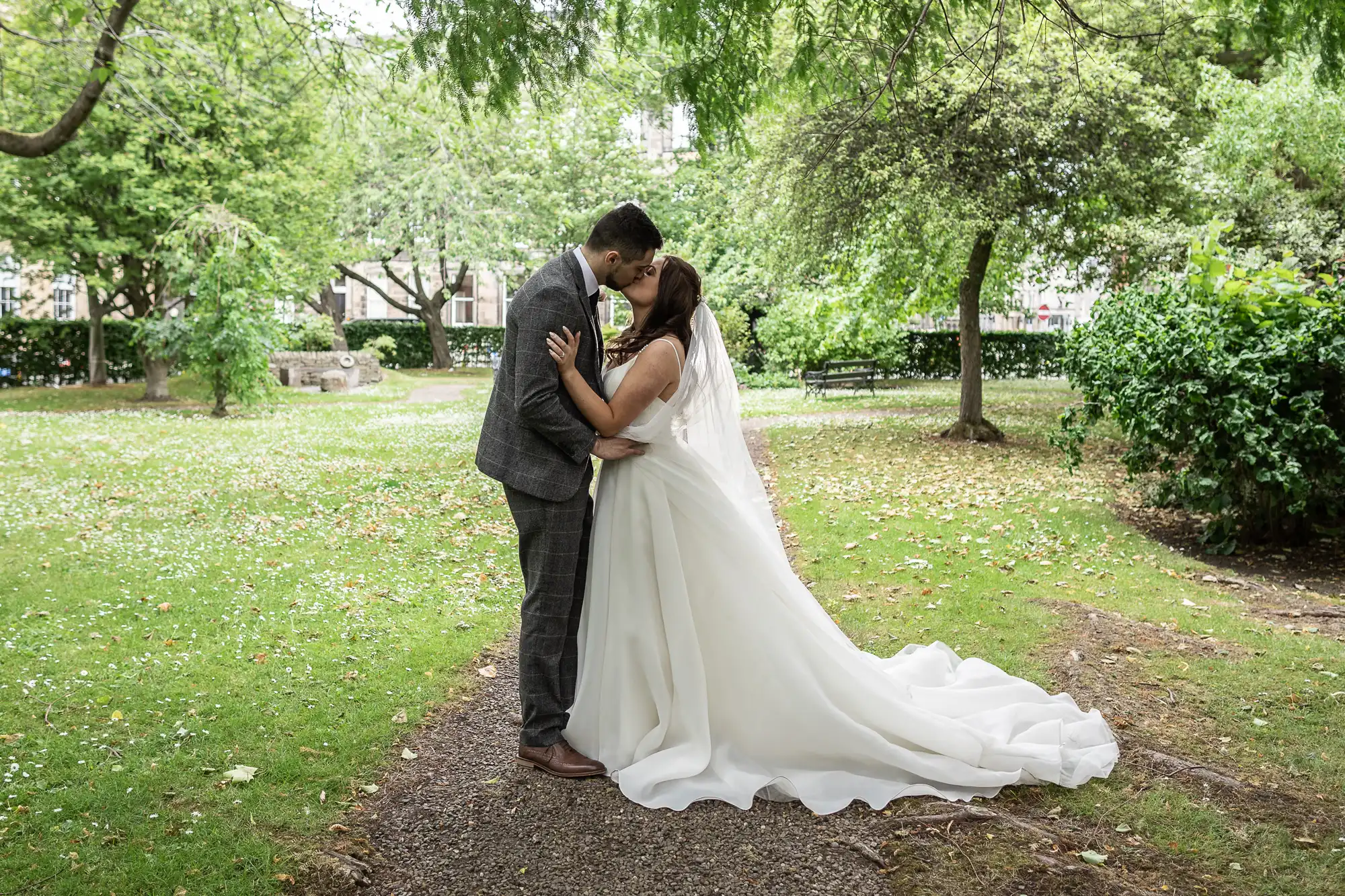 A bride and groom kissing in a lush garden, the bride in a long white dress with a train, and the groom in a gray suit.