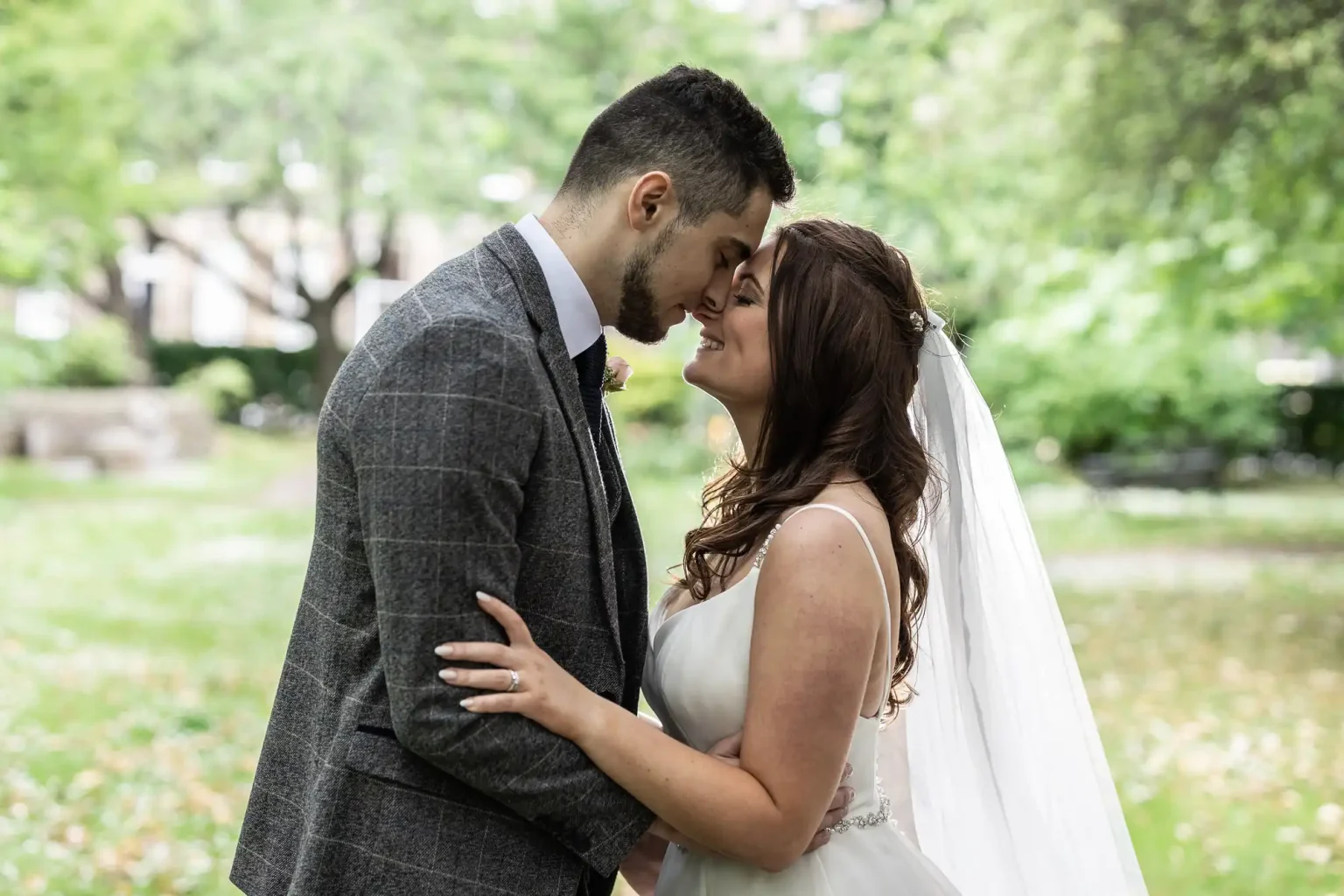 A newlywed couple touching foreheads lovingly, with the groom in a gray suit and the bride in a white gown, standing in a lush park.