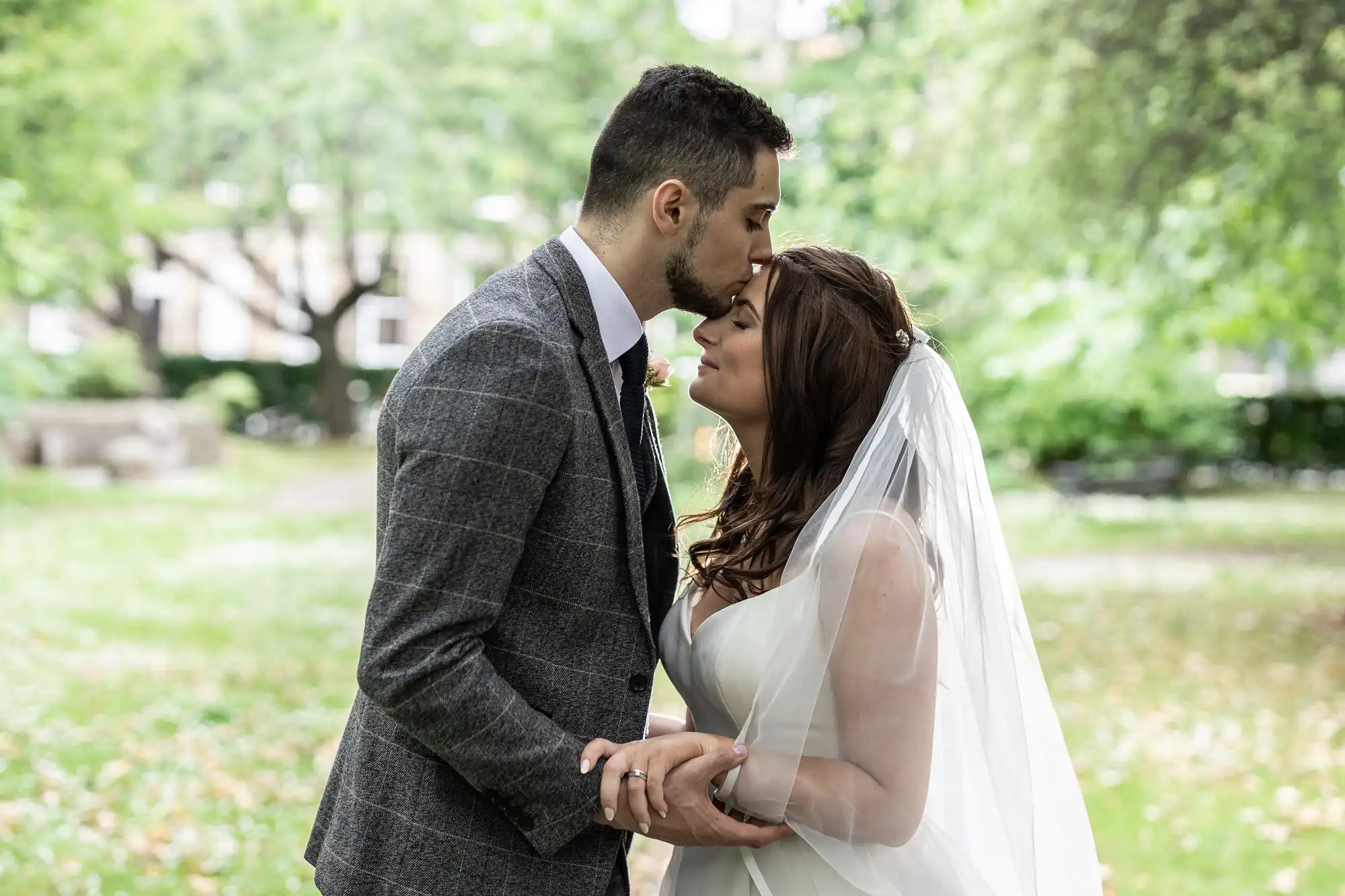 A bride and groom softly touching foreheads in a lush park, both dressed in wedding attire.