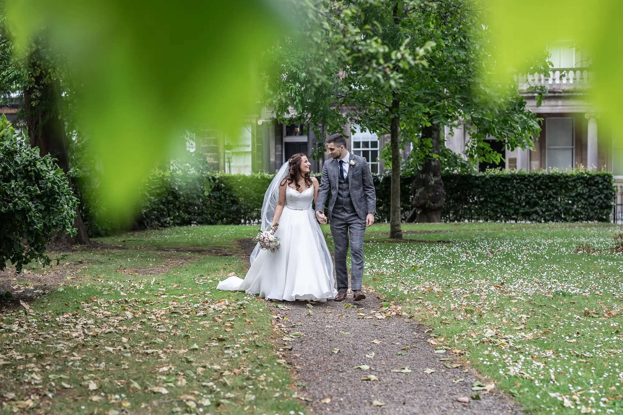 A bride and groom walking on a garden path, partially framed by green leaves, with the bride holding a bouquet.