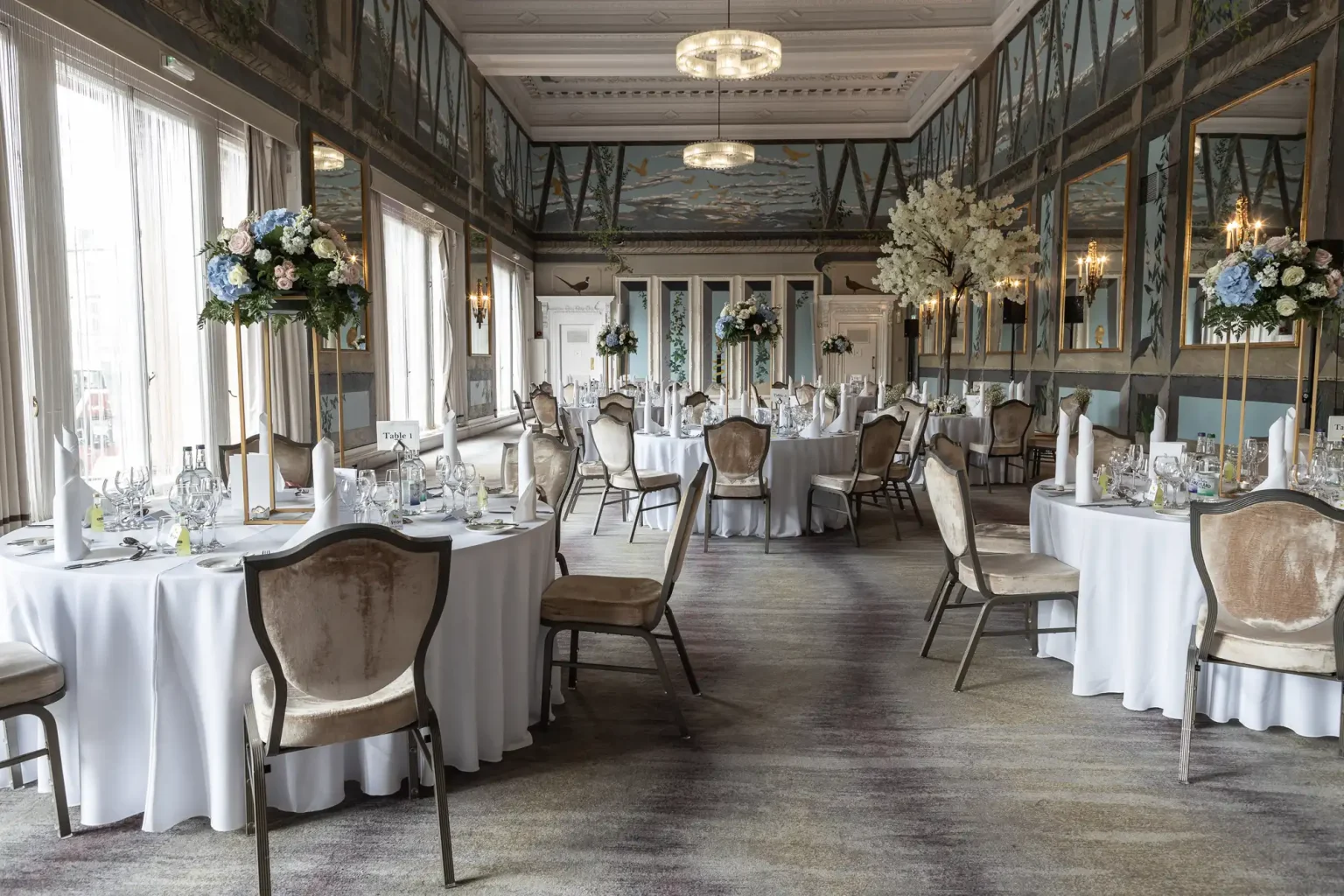 An elegant dining room set up for a formal event with round tables adorned with white tablecloths, floral centerpieces, and surrounded by vintage chairs.