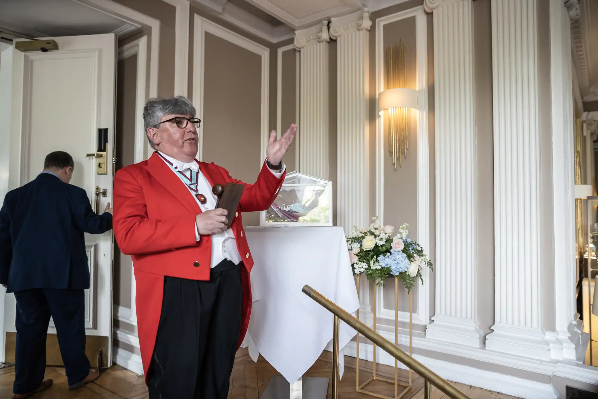 An older man with gray hair, wearing a red jacket and glasses, gestures while speaking in an elegant room with columns and chandeliers. Another man is partially visible in the background.