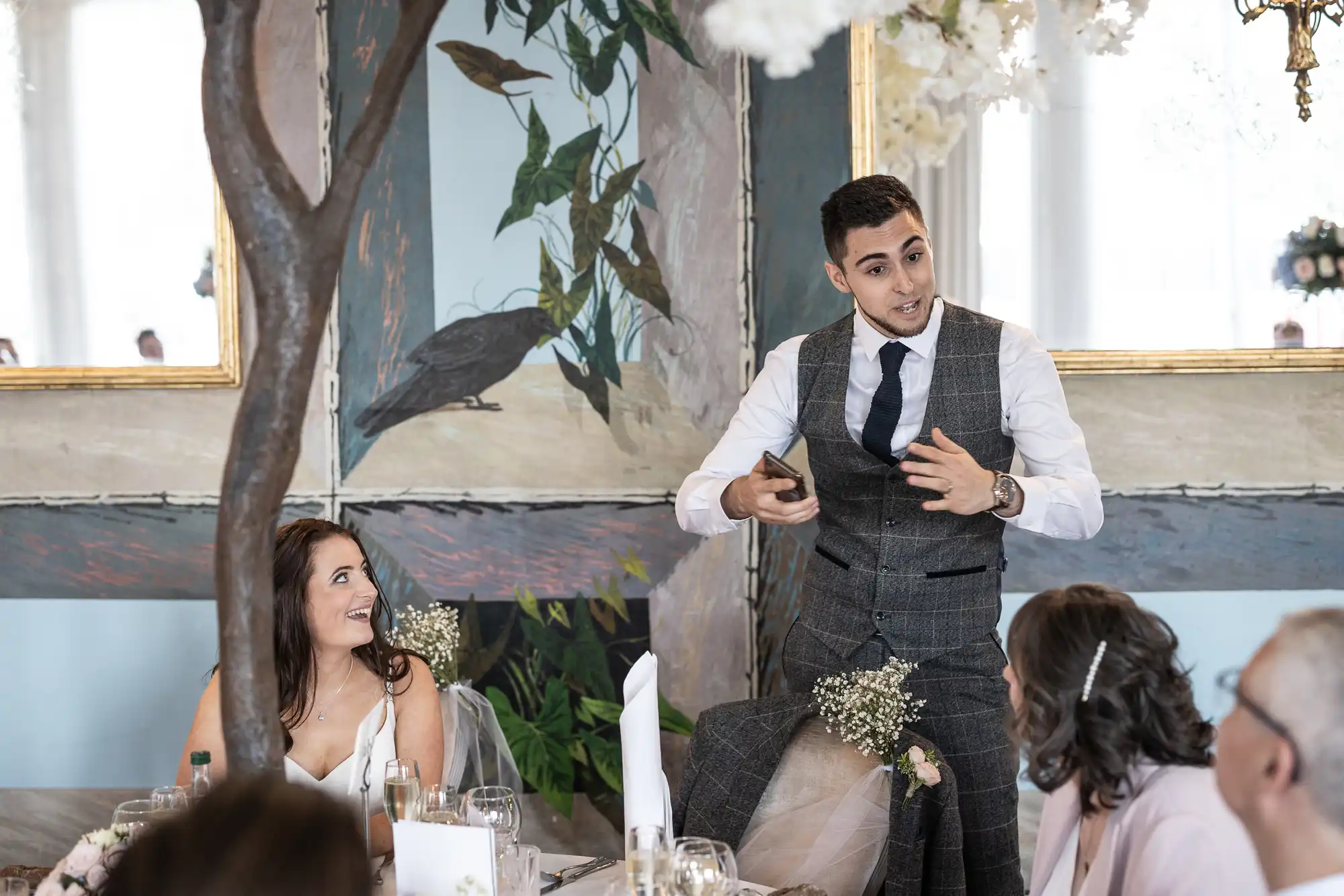 Groom speech stands speaking animatedly at a wedding, with guests looking on and smiling in a room with tree and bird murals.