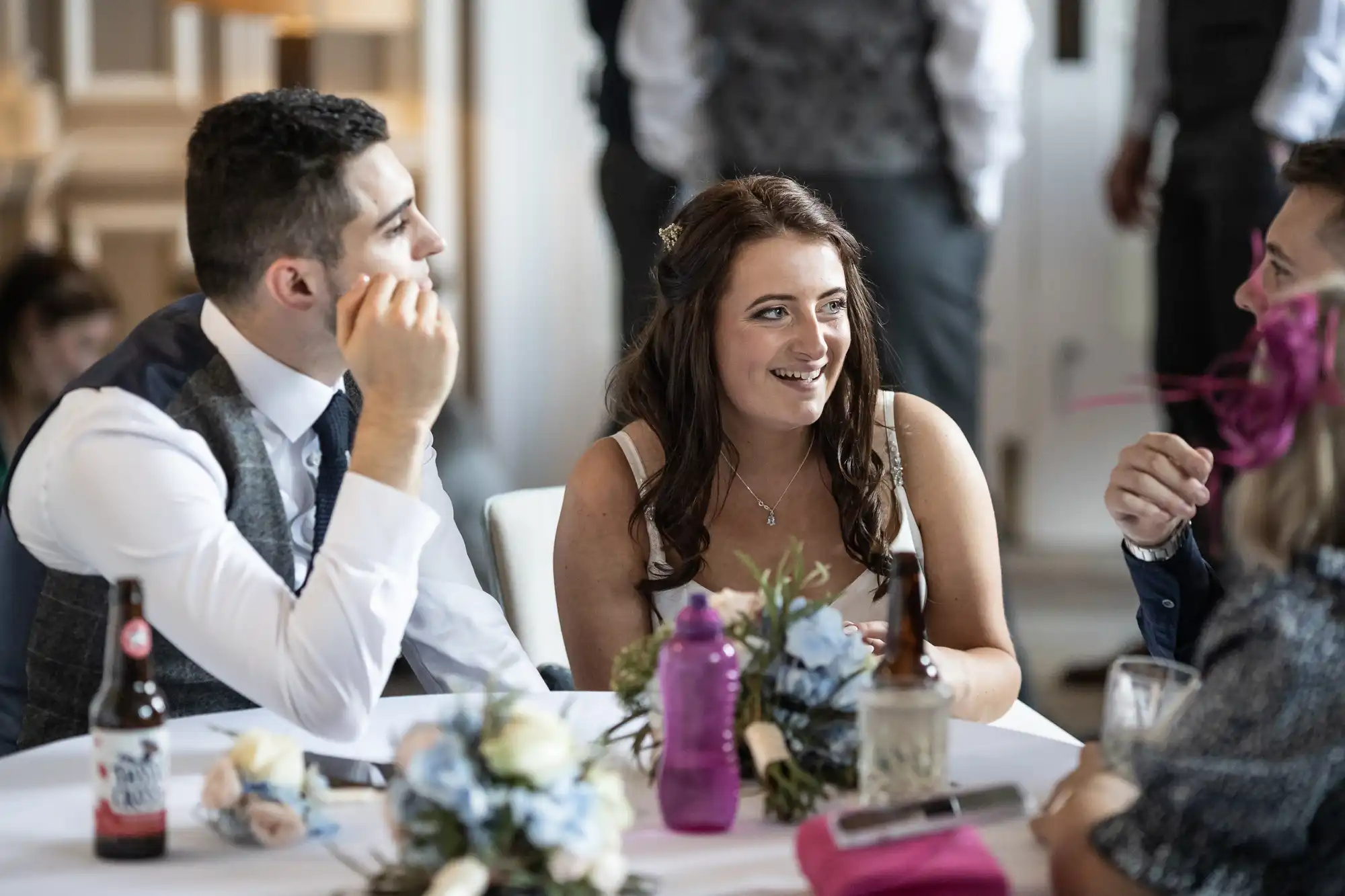 A woman and two men engaged in conversation at a wedding reception table, with refreshments and floral decorations visible.