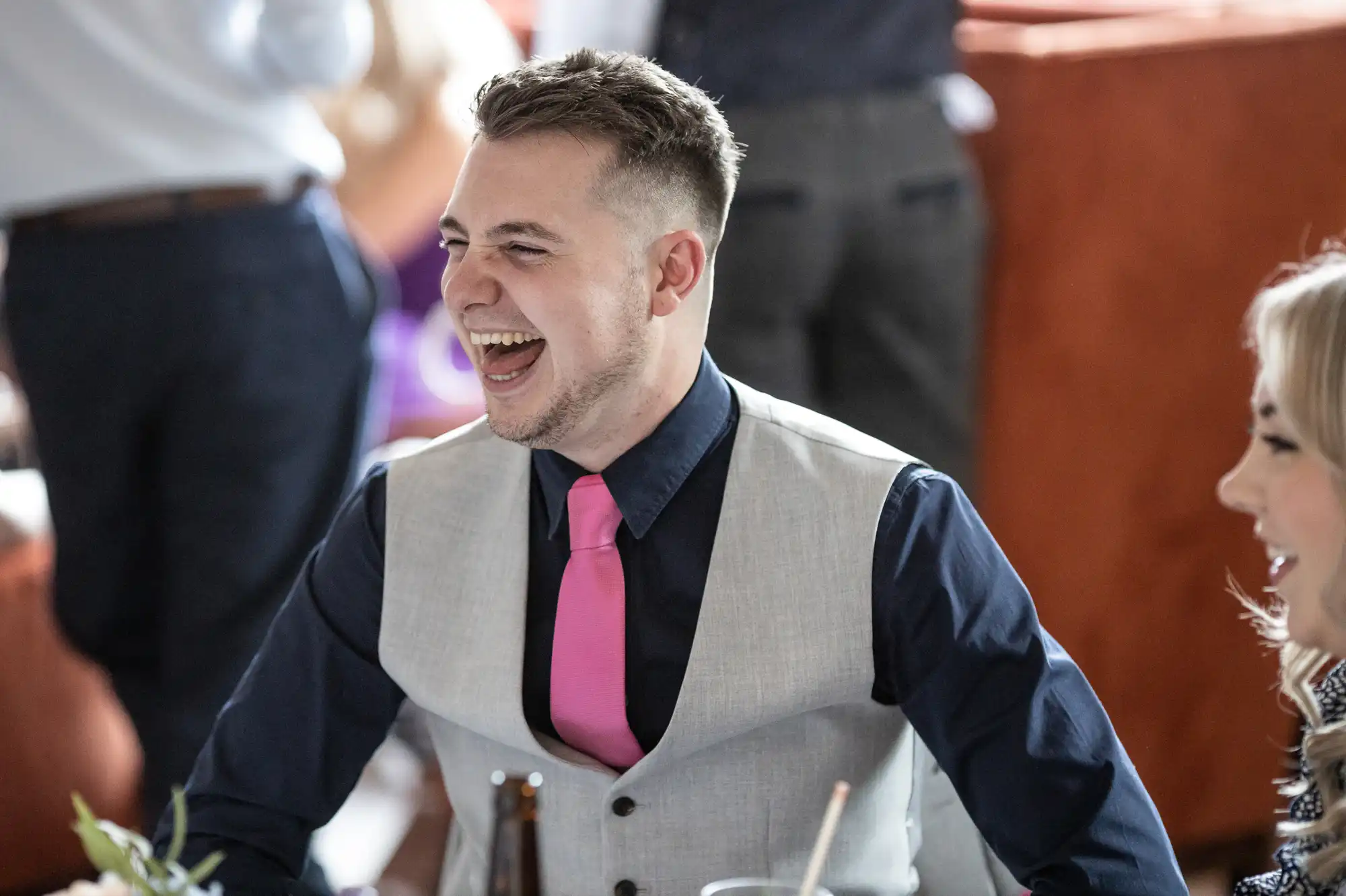 A man in a formal gray vest and pink tie laughing joyfully at a social event, seated at a table with visible beverages.