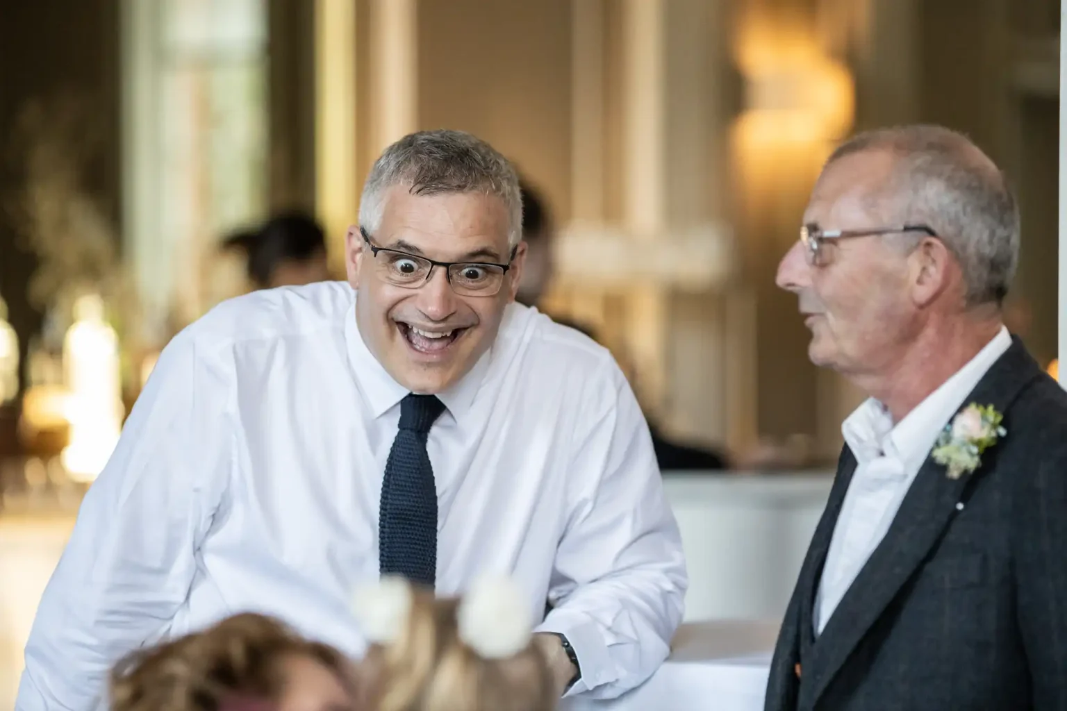 Two older men in formal wear having a joyful conversation at an event, with one man leaning forward smiling broadly.