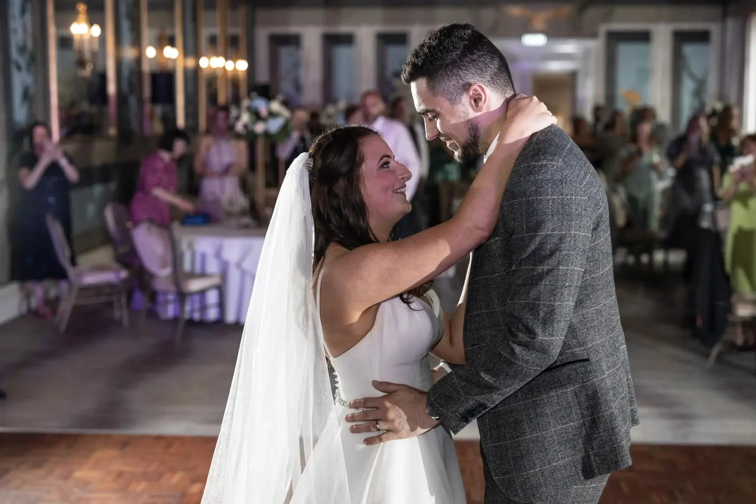 A bride and groom share a tender moment on the dance floor at their wedding reception, with guests watching in the background.