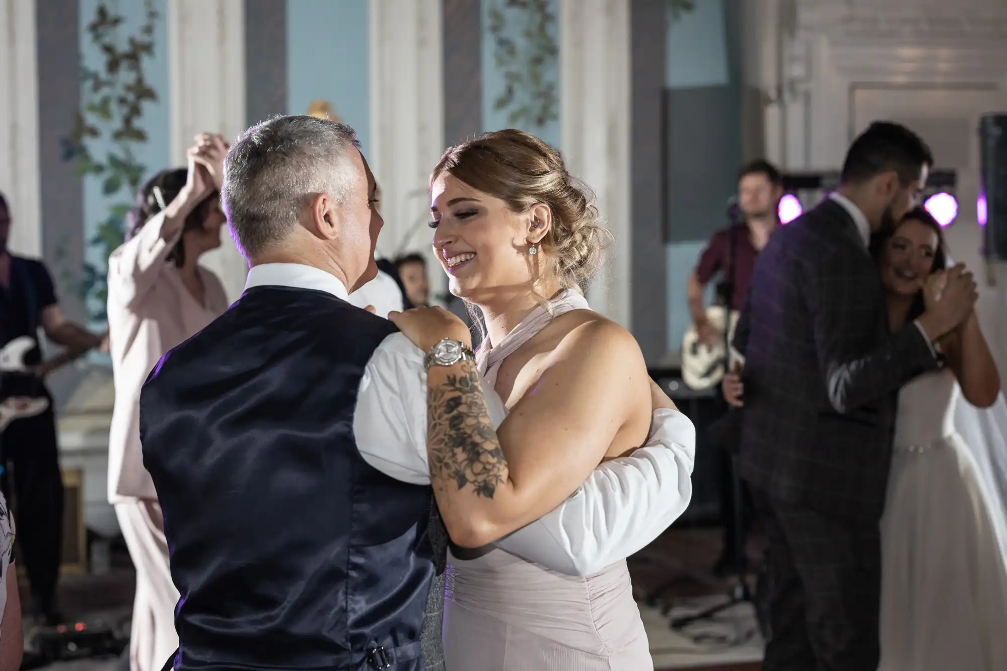 A bridesmaid and her partner share a warm, smiling dance at a wedding reception, surrounded by dancing guests in an elegantly lit hall.