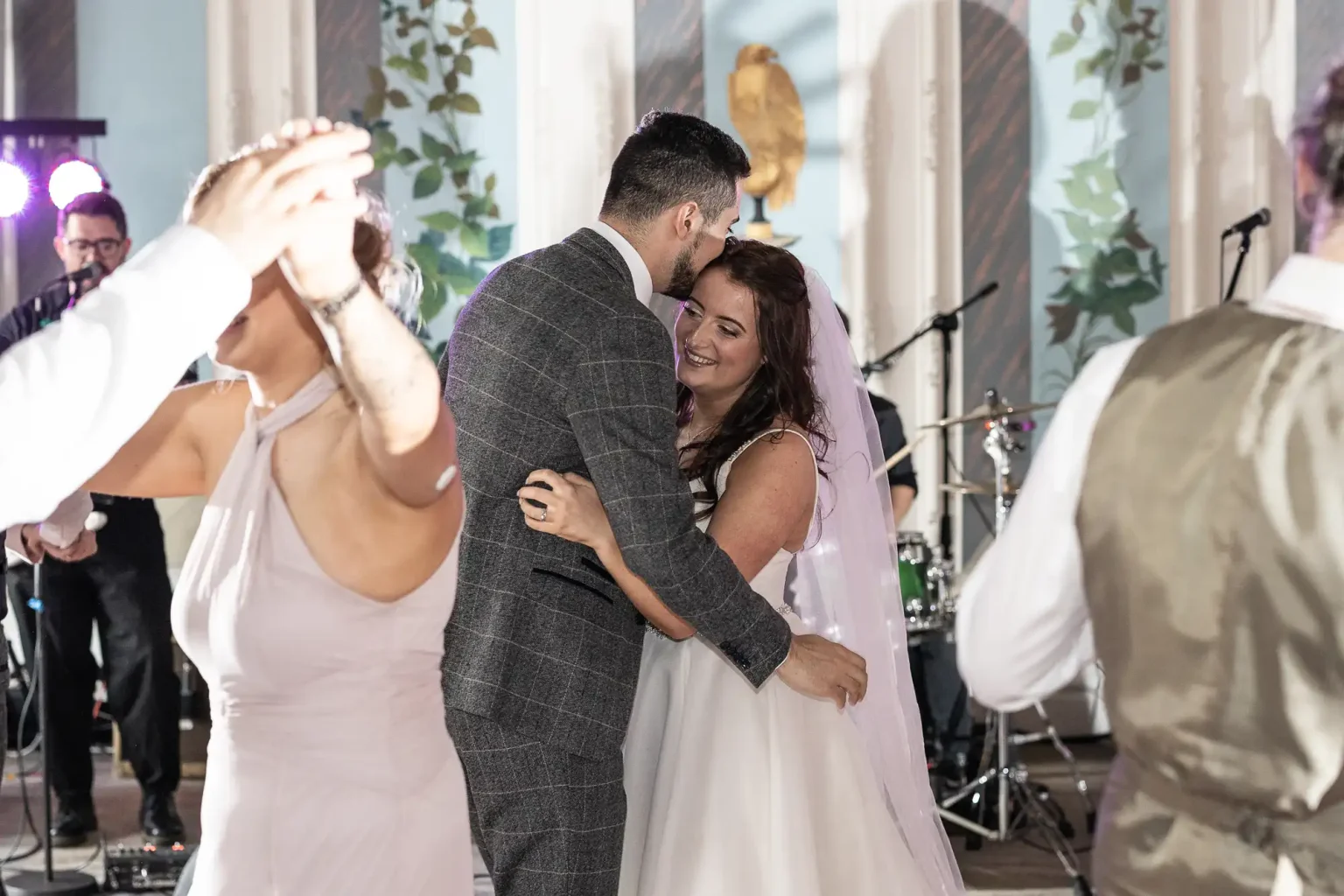 A bride and groom dancing closely at their wedding reception, surrounded by guests and a live band in the background.