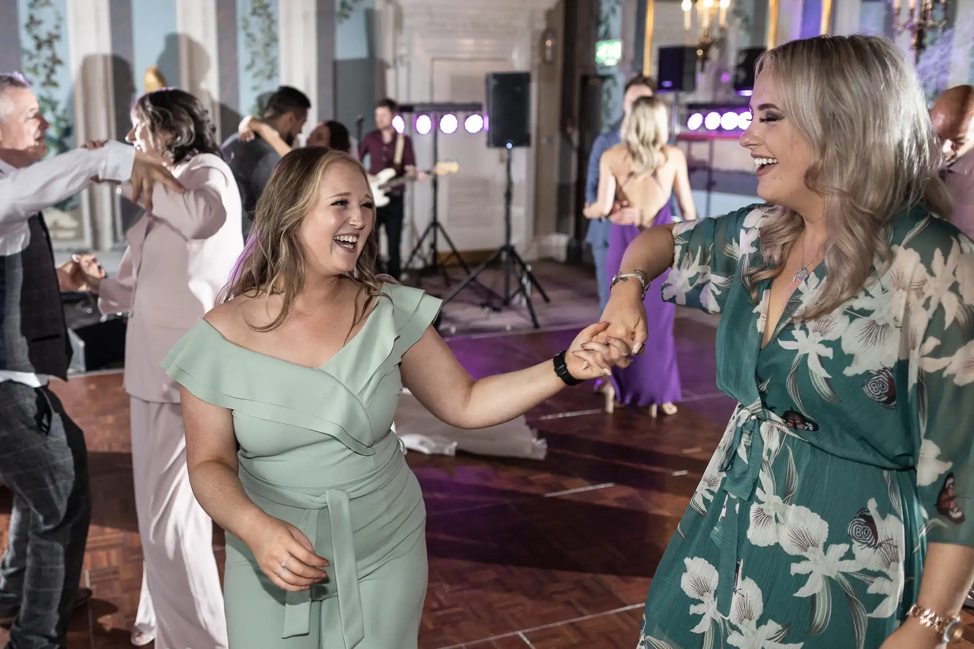 Two women joyfully dancing and holding hands at a lively indoor wedding reception, with other guests dancing and a live band in the background.