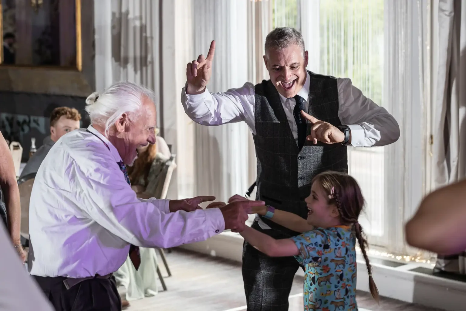 A young girl and two older men smiling while playing a game indoors, one man gestures excitedly with a raised finger.