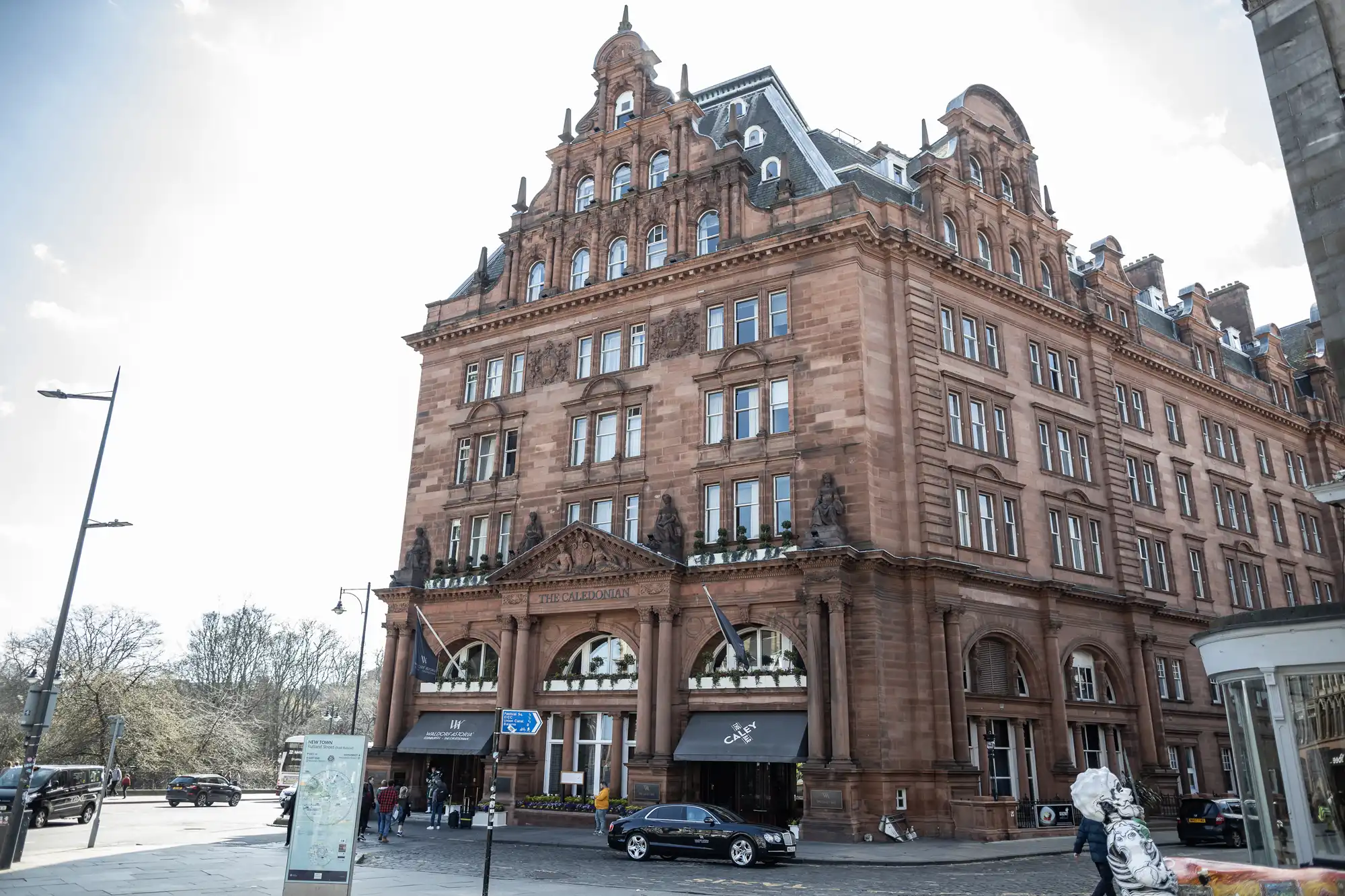 A grand, multi-story red-brick building with arched windows and an elaborate facade, located on a street corner with cars and pedestrians nearby.