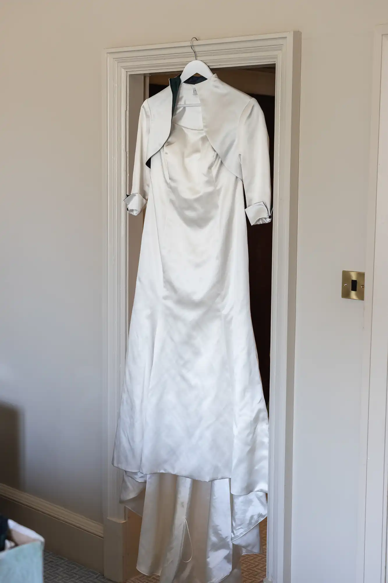 A white satin wedding dress and matching bolero jacket hang on a wooden hanger in a doorway.