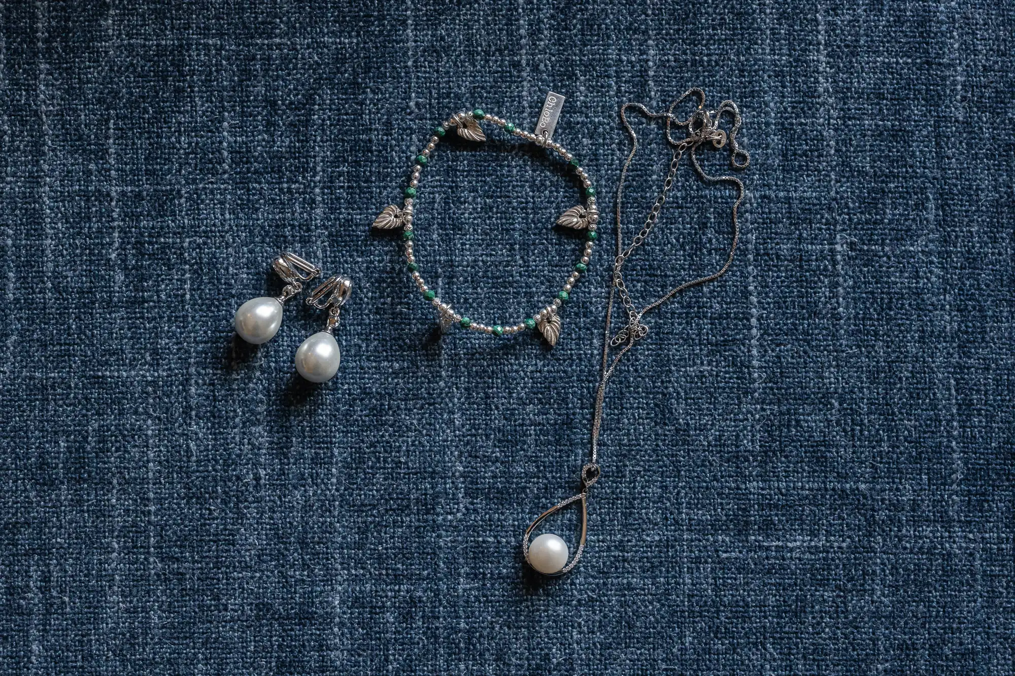 A pair of pearl earrings, a beaded bracelet with a leaf design, and a pearl necklace with a chain are displayed on a textured blue surface.