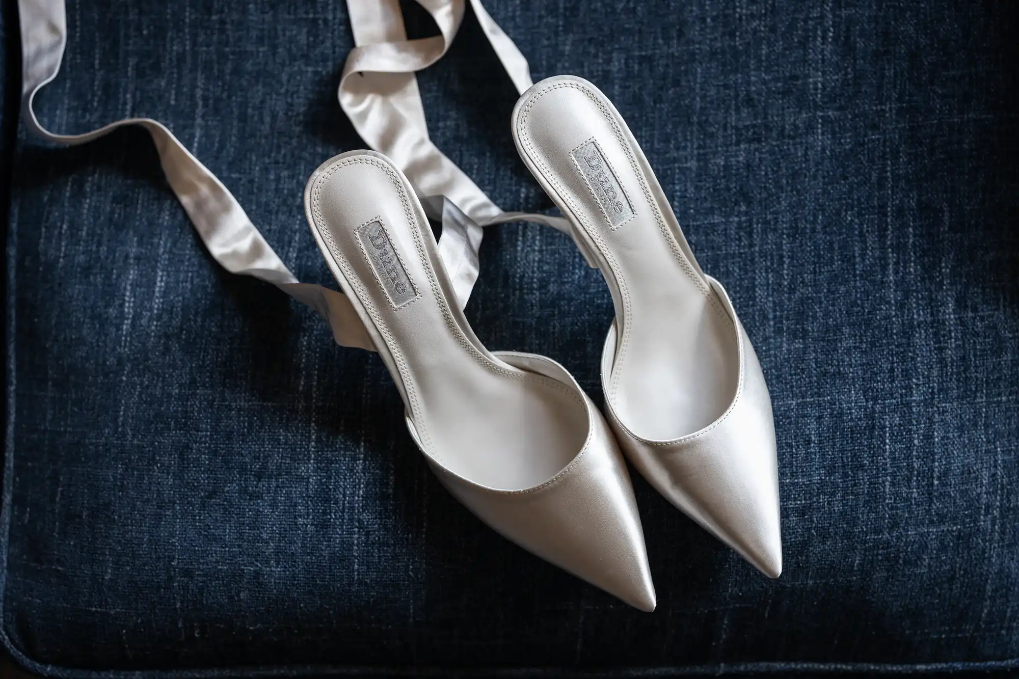 A pair of satin pointed-toe shoes with ribbon straps on a dark blue fabric background.