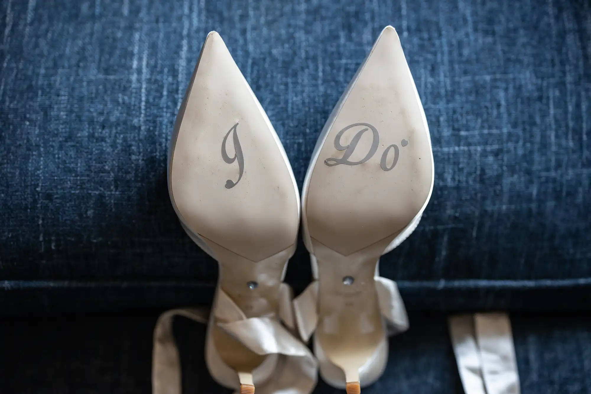 A close-up of the soles of a pair of white high-heeled shoes with "I Do" written on them, placed on a dark blue fabric background.