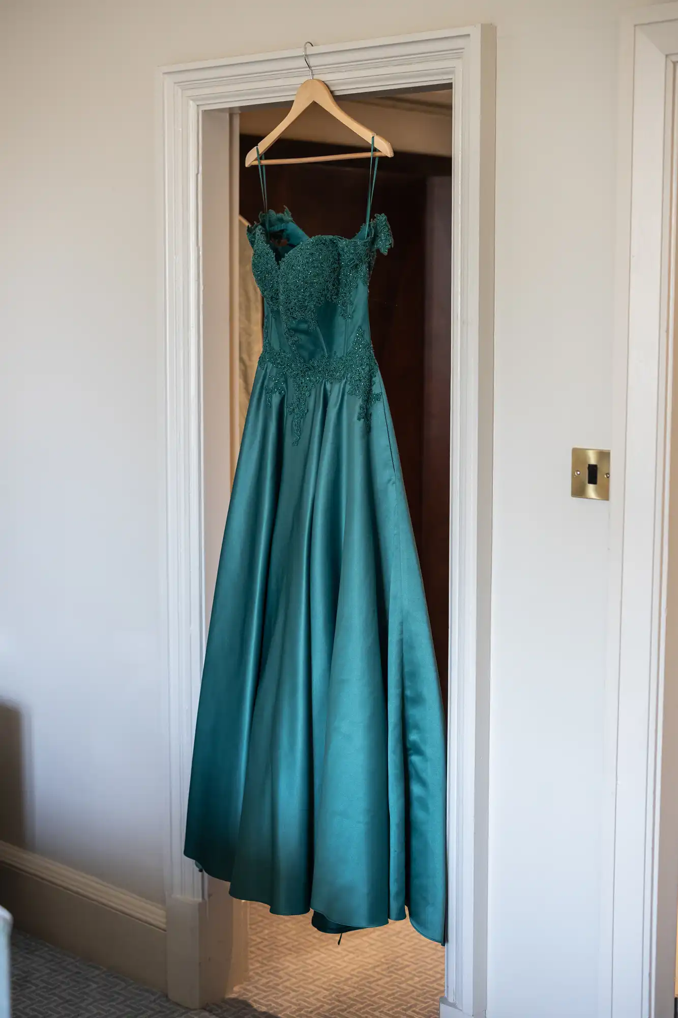 A teal formal gown with lace detailing and spaghetti straps hangs on a wooden hanger in a doorway.