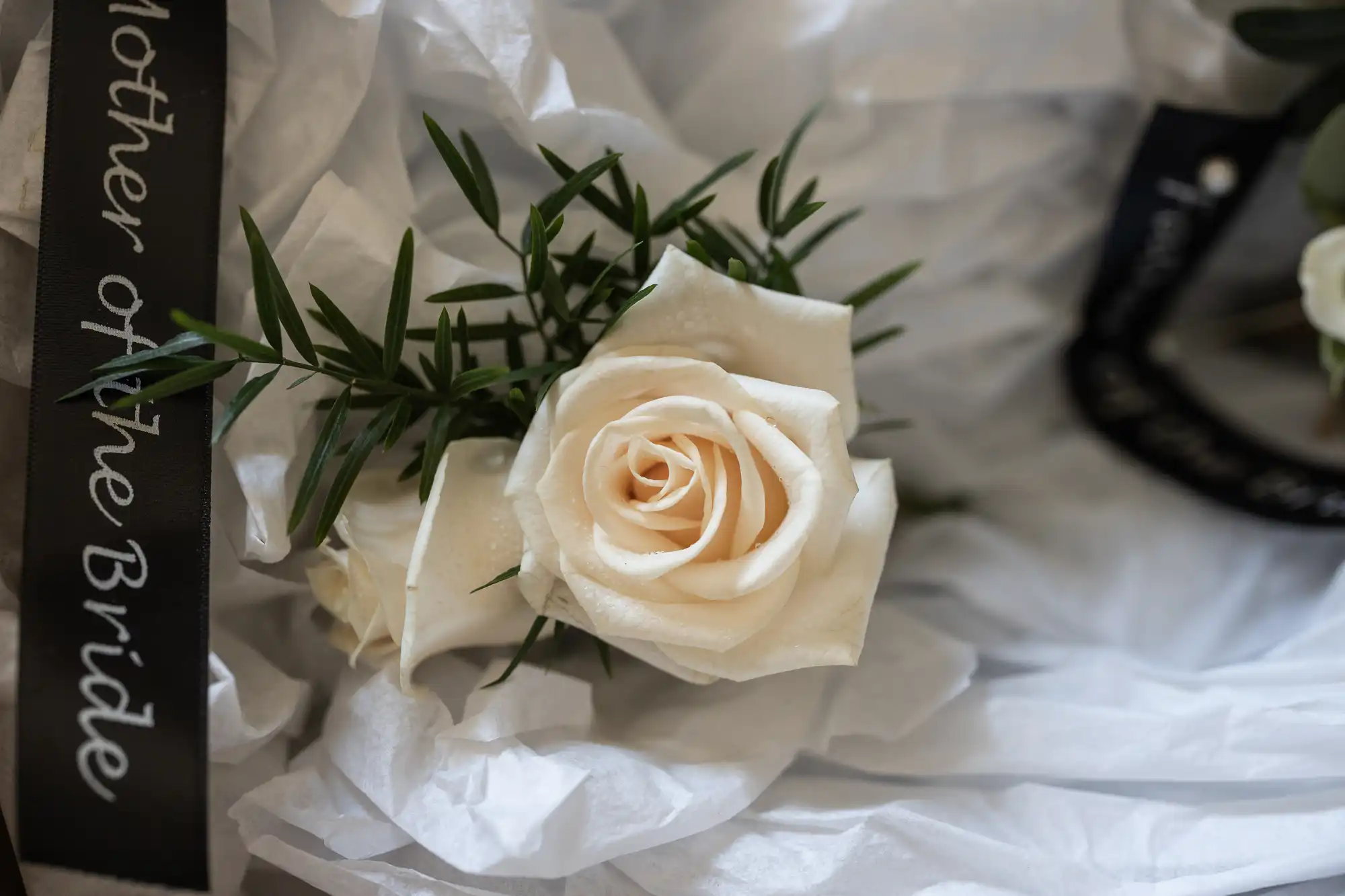 A white rose corsage labeled "Mother of the Bride" rests on white tissue paper with greenery accents.