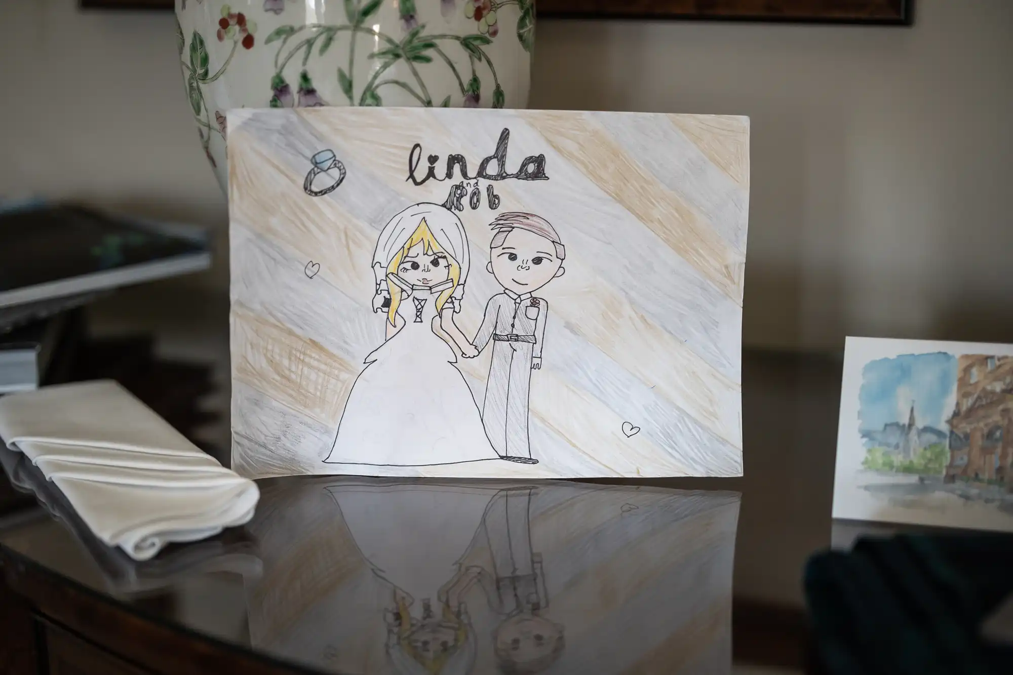 A child’s drawing of a bride and groom, labeled "Linda and Rib," displayed on a table with a folded white napkin and a potted plant in the background.