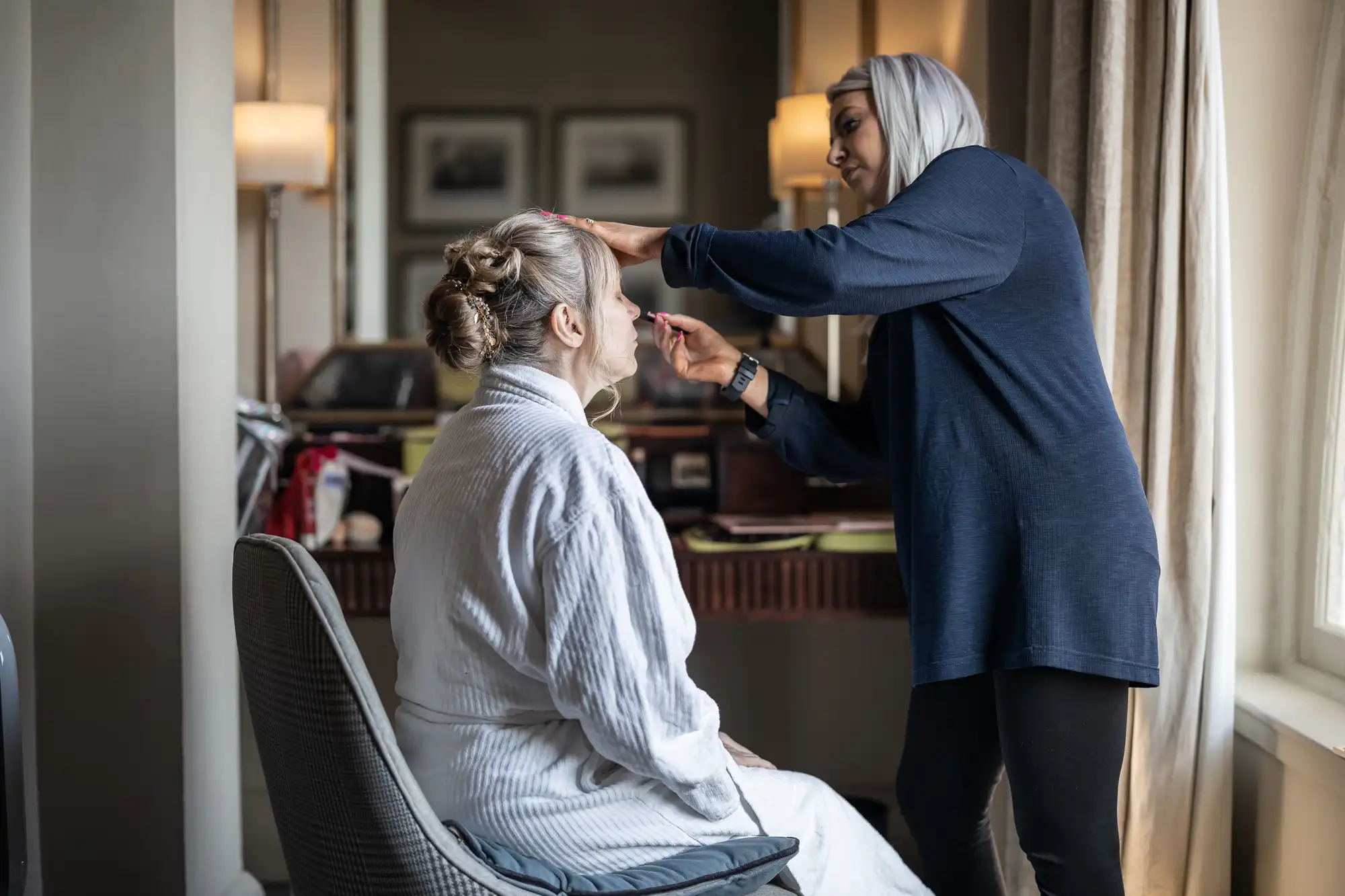 A makeup artist is applying makeup to a woman sitting in a chair in a well-lit room. The woman is wearing a white robe and has her hair styled in an updo.