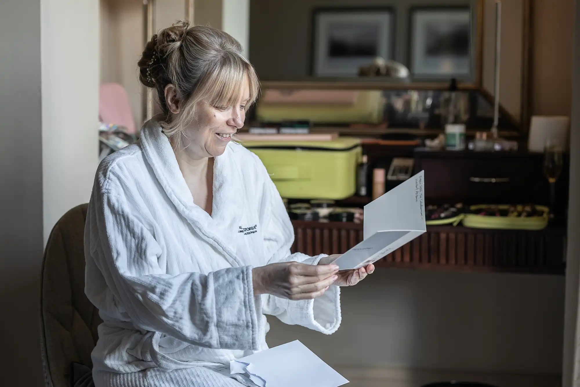 A woman in a white bathrobe sits at a dresser and reads a card with a smile. The dresser is organized with various makeup items and personal care products.