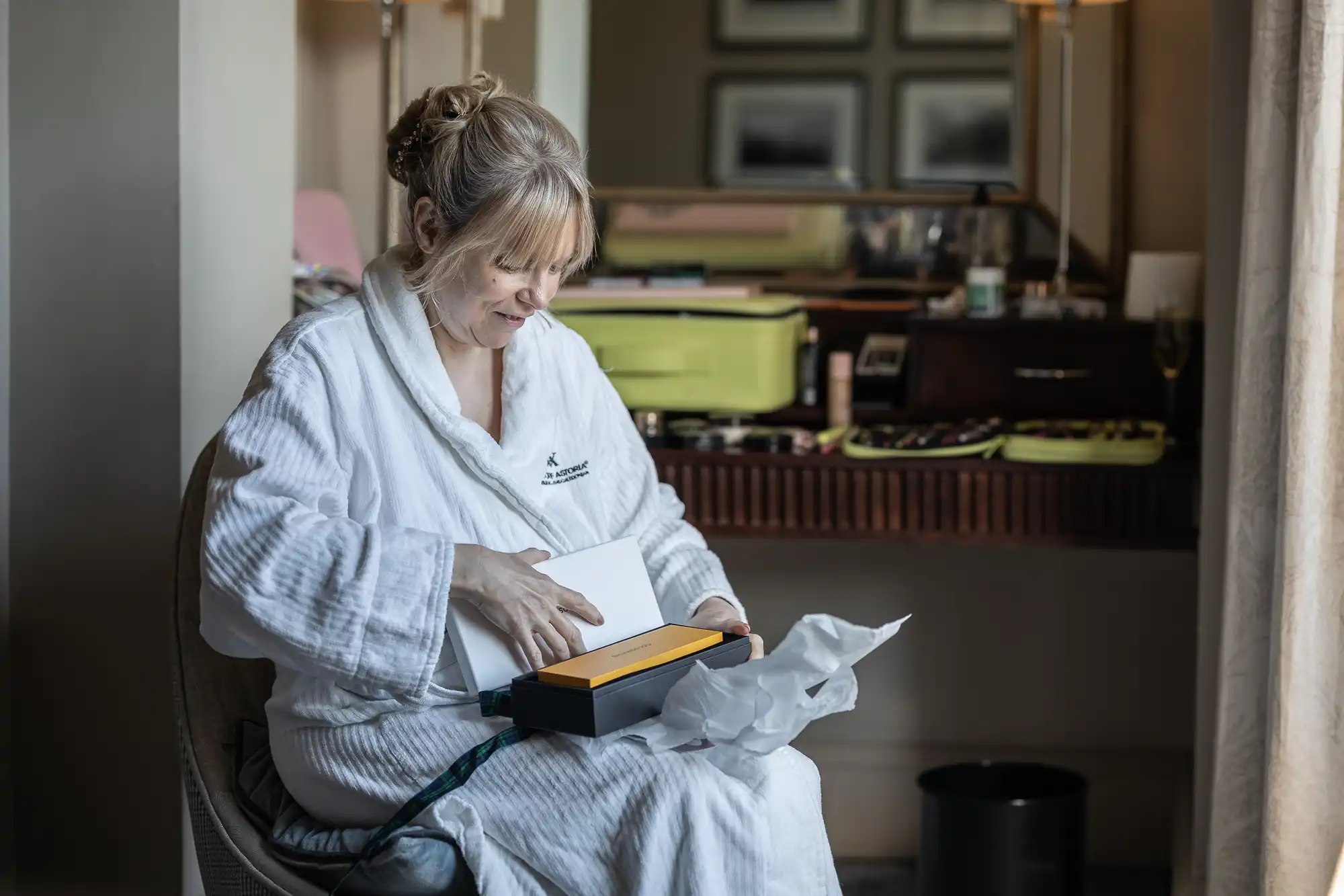 A woman in a white robe sits on a chair, opening a box. The box has tissue paper and a yellow item inside. The background shows a table with various objects and a mirror.