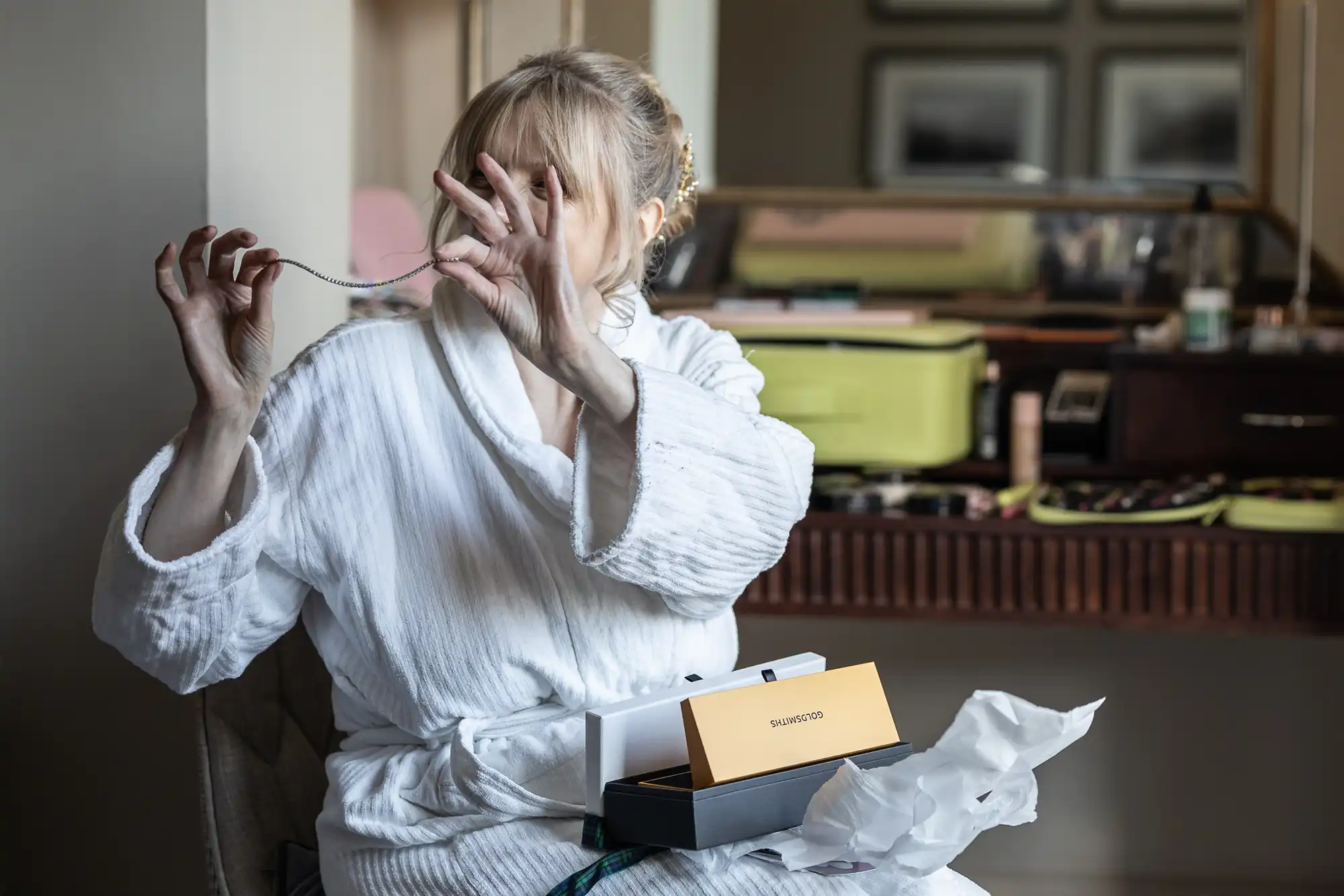 Person in a white robe sitting and examining a necklace from a SUENOSO box. A dresser with various items is visible in the background.