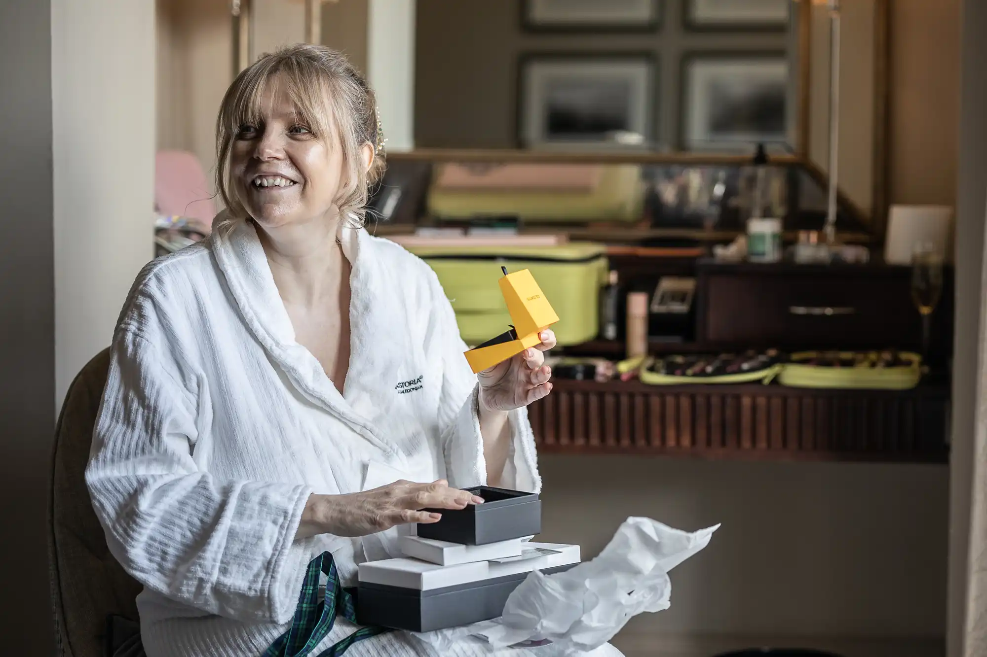 A woman in a white bathrobe is smiling while holding a small yellow box, sitting in a room with various items on a table in the background.