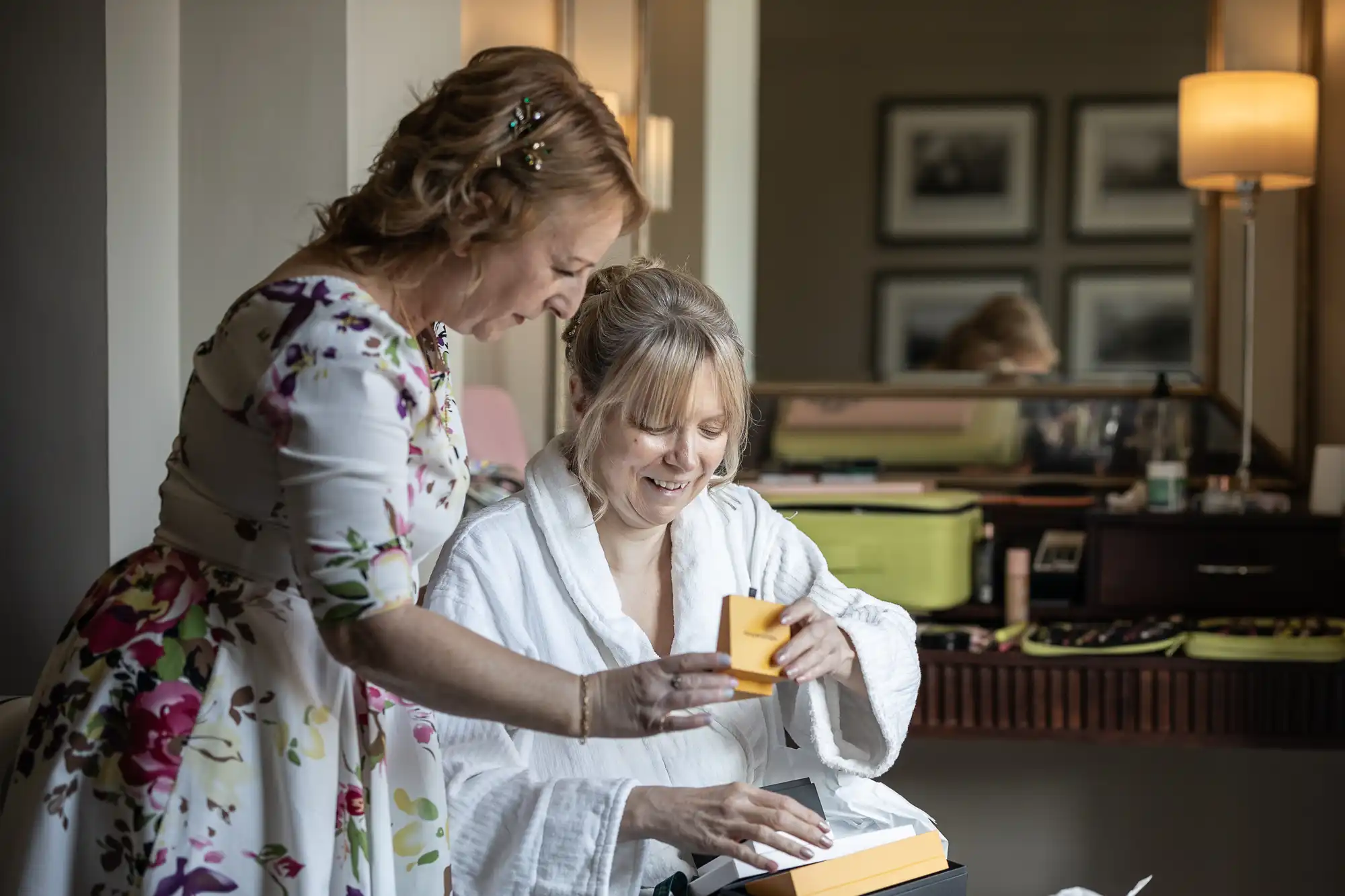 Two women in a room. One is wearing a floral dress, the other is in a bathrobe, holding a small yellow box. They are smiling and looking at the contents of the box.