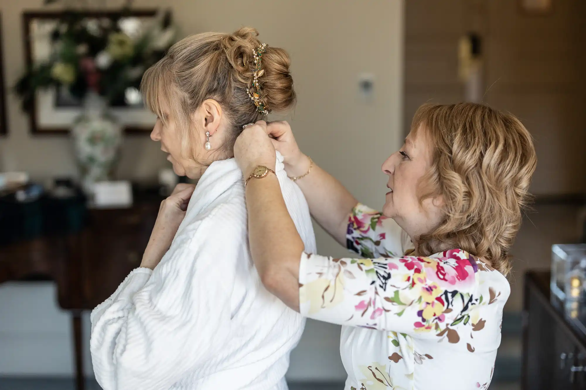 Two women are in a room. One woman, wearing a floral dress, is adjusting the back of a white garment on the other woman, who has her hair styled in an updo.