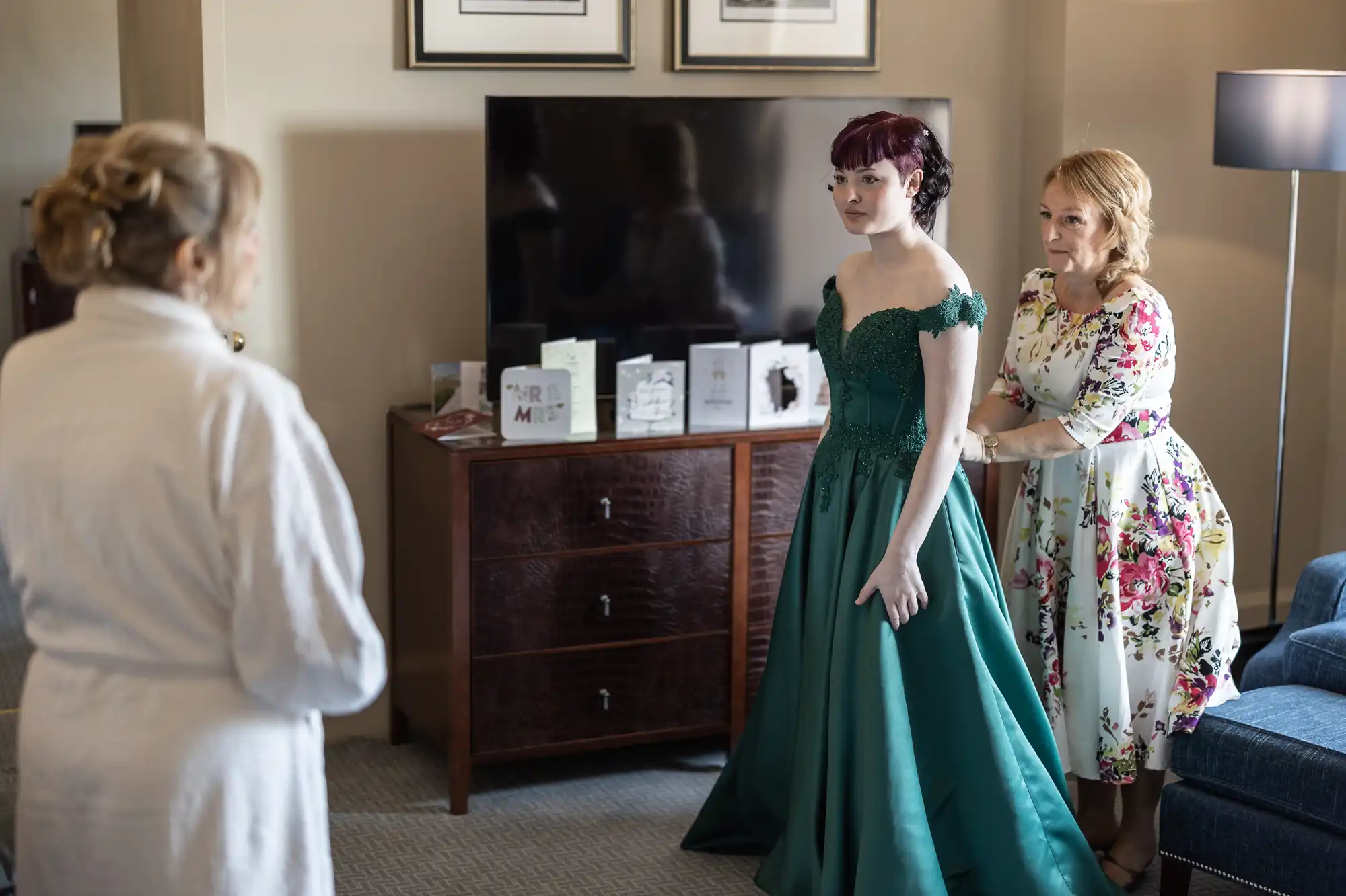 Two women help a third woman in a green gown adjust her dress in a living room setting. There are cards displayed on a wooden dresser in the background.