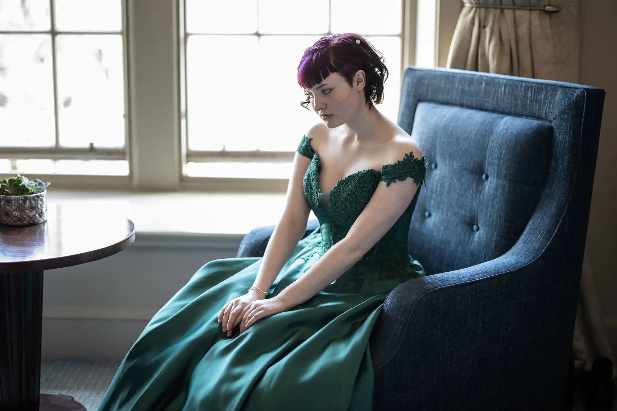 A woman in a green dress sits on a blue chair near a window, looking pensive.