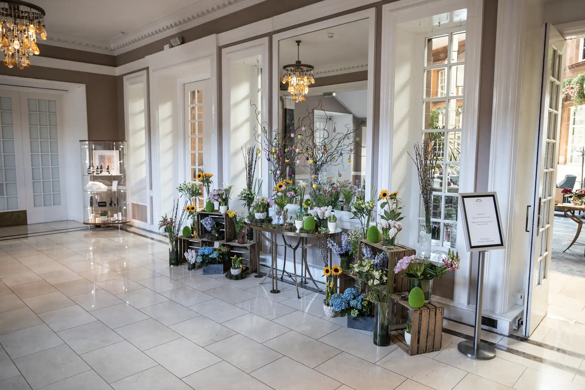 A bright room with tiled floor features a display of various flowers, including sunflowers and hydrangeas, arranged on tables and in crates in front of large mirrors and windows. A sign stands nearby.