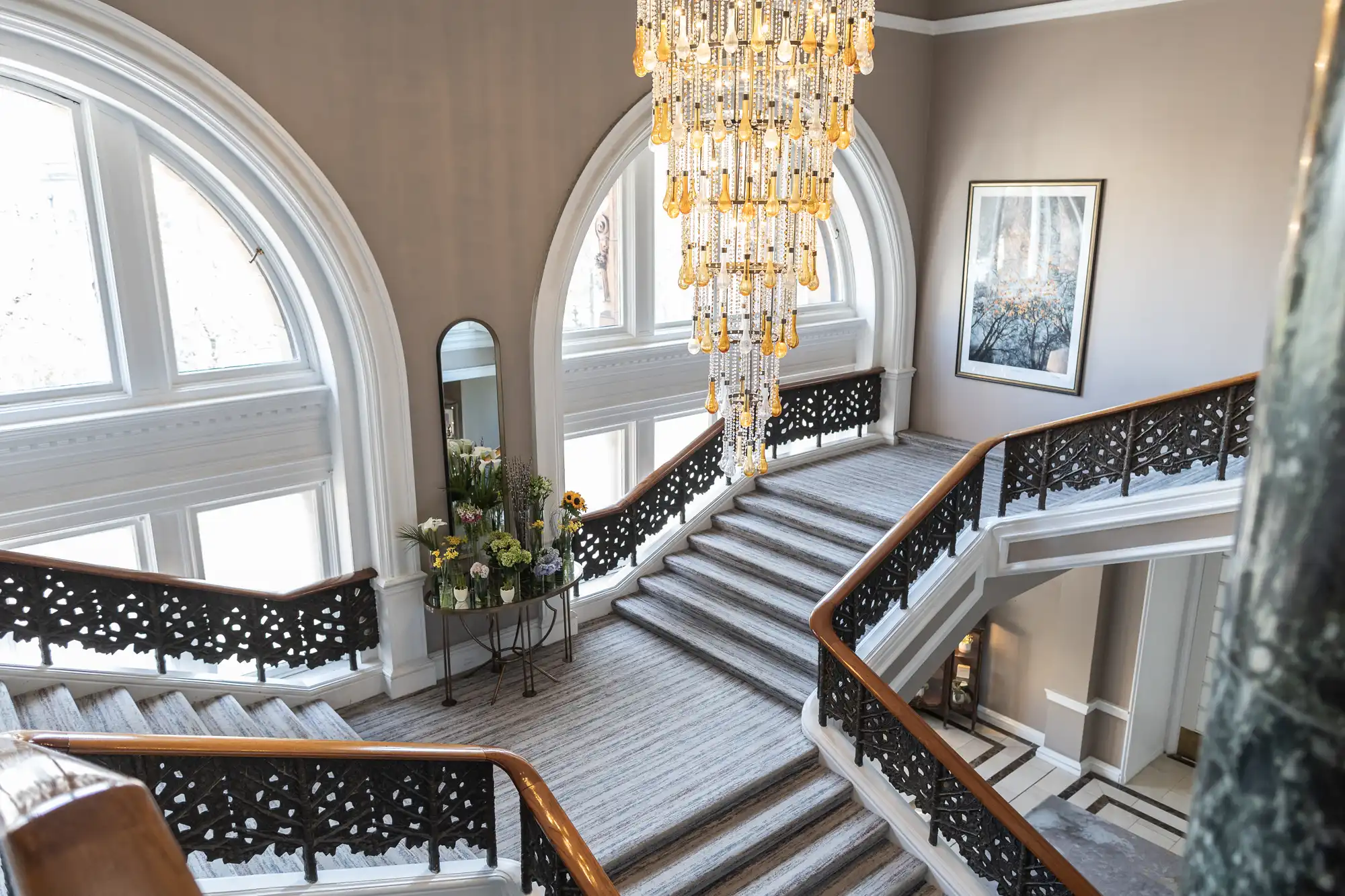 Elegant staircase with ornate black railing, fluffy grey carpet, and a large crystal chandelier. Decor includes a tall mirror, wall art, and a floral arrangement. Arched windows light the area.