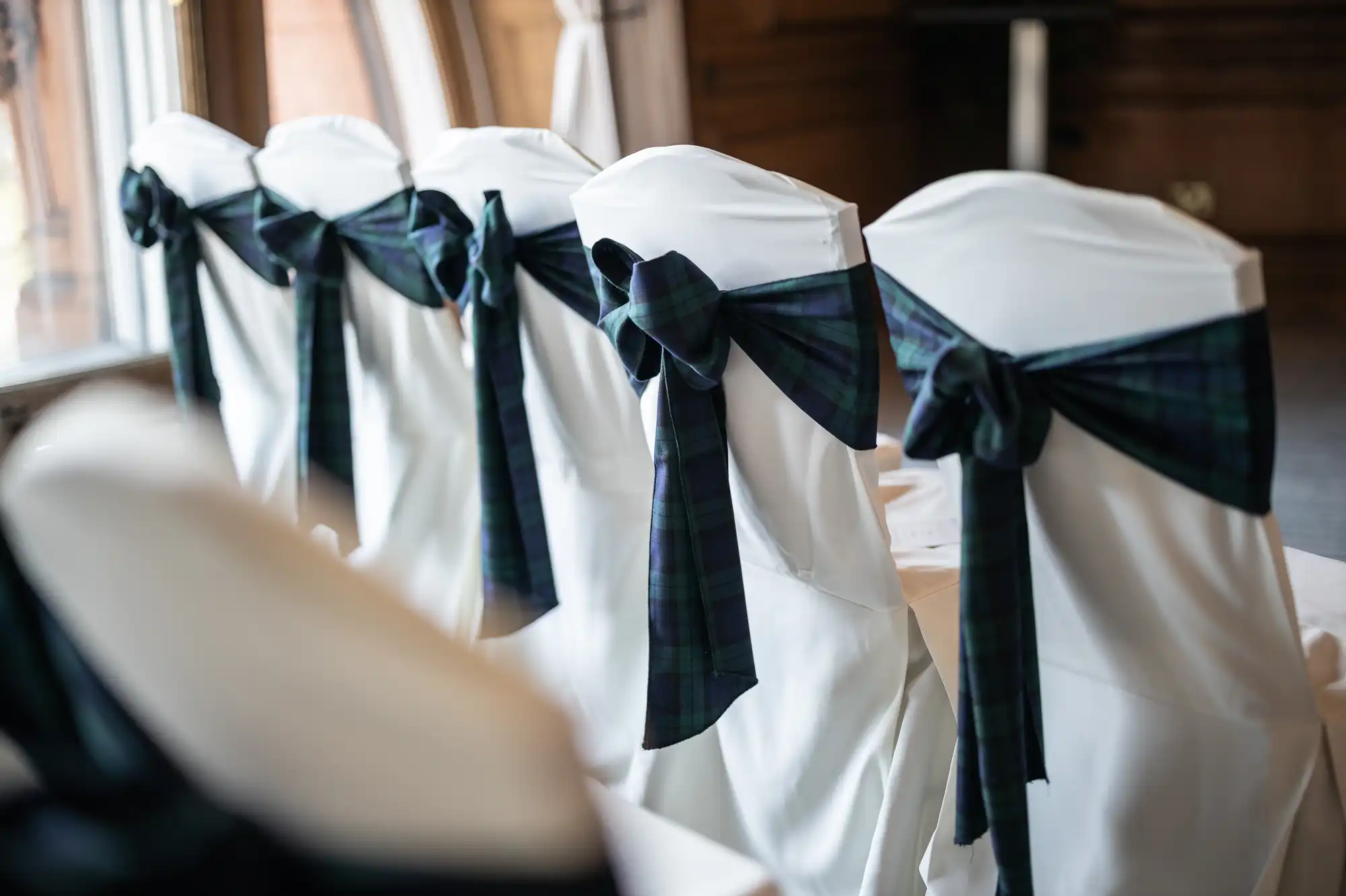 Five white chairs with dark plaid sashes tied in bows along a row by a window.