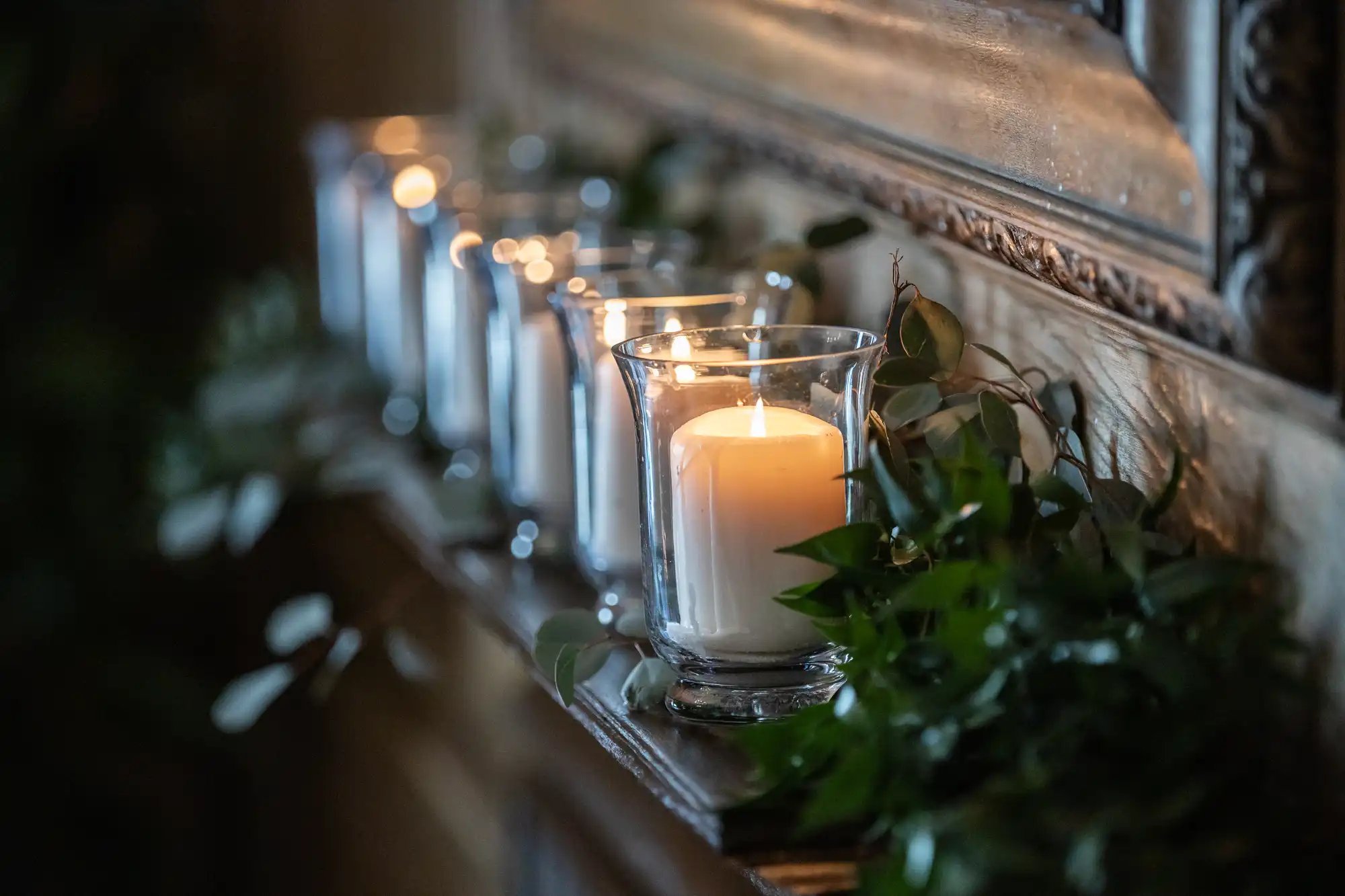 A row of lit candles in glass holders placed on a wooden shelf, surrounded by green foliage.