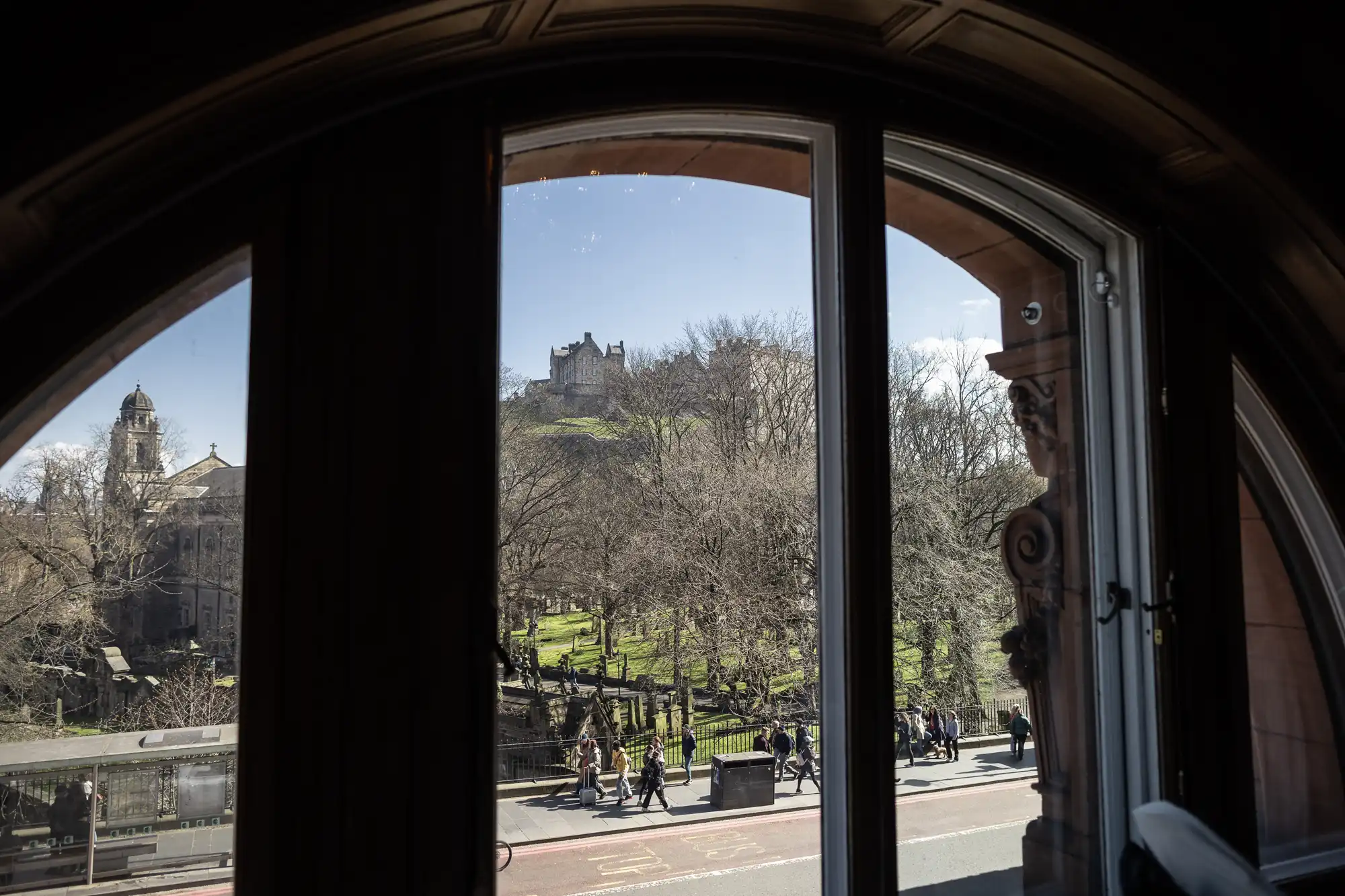 View through an arched window showing a busy street with pedestrians and a distant hilltop castle under a clear sky.