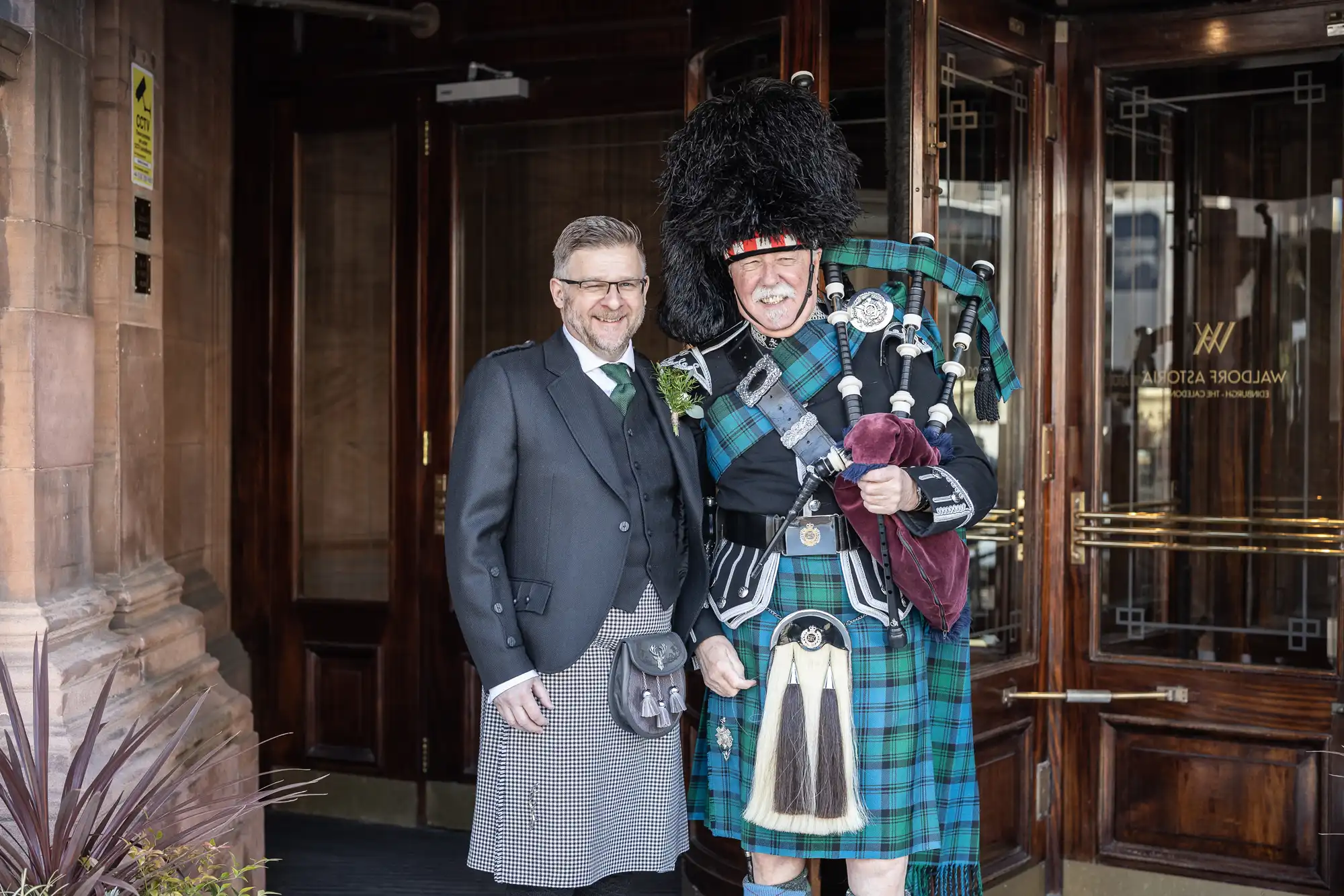 Two men stand at an entrance. One wears a kilt and waistcoat, while the other wears a traditional Scottish attire with a bagpipe.