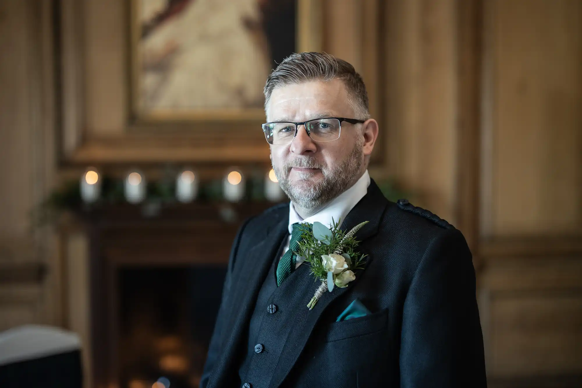 A man with glasses, wearing a formal suit with a boutonniere, stands indoors in front of a wooden wall with a framed photo.