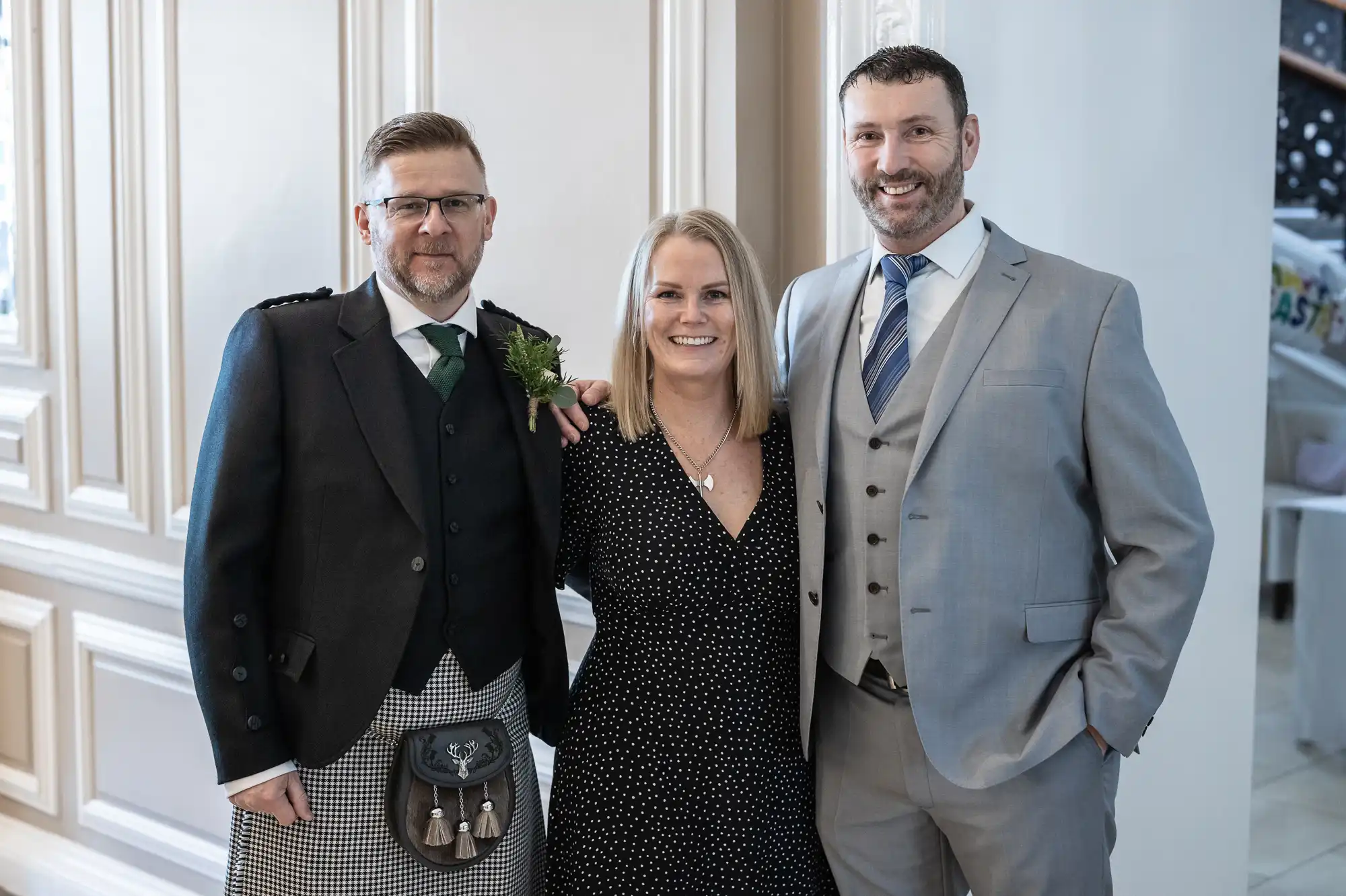 Three people are standing indoors, smiling at the camera. The man on the left wears a kilt and sporran, while the woman in the center and the man on the right wear formal attire.