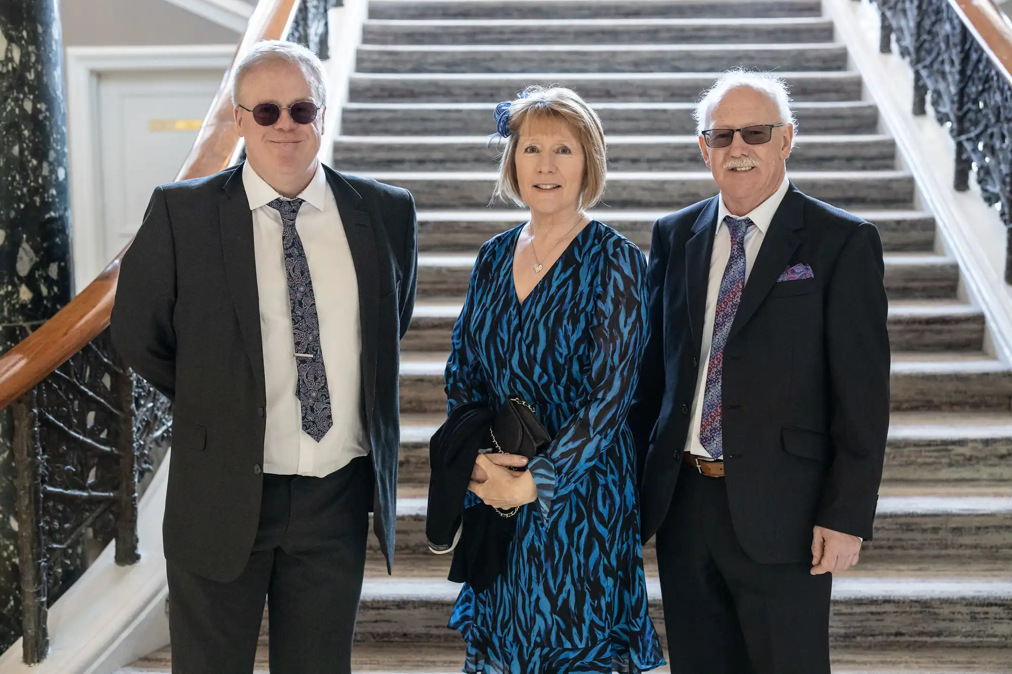 Three individuals are dressed in formal attire, standing in front of a staircase with elegant railings. The two men are wearing suits, and the woman is wearing a blue patterned dress.