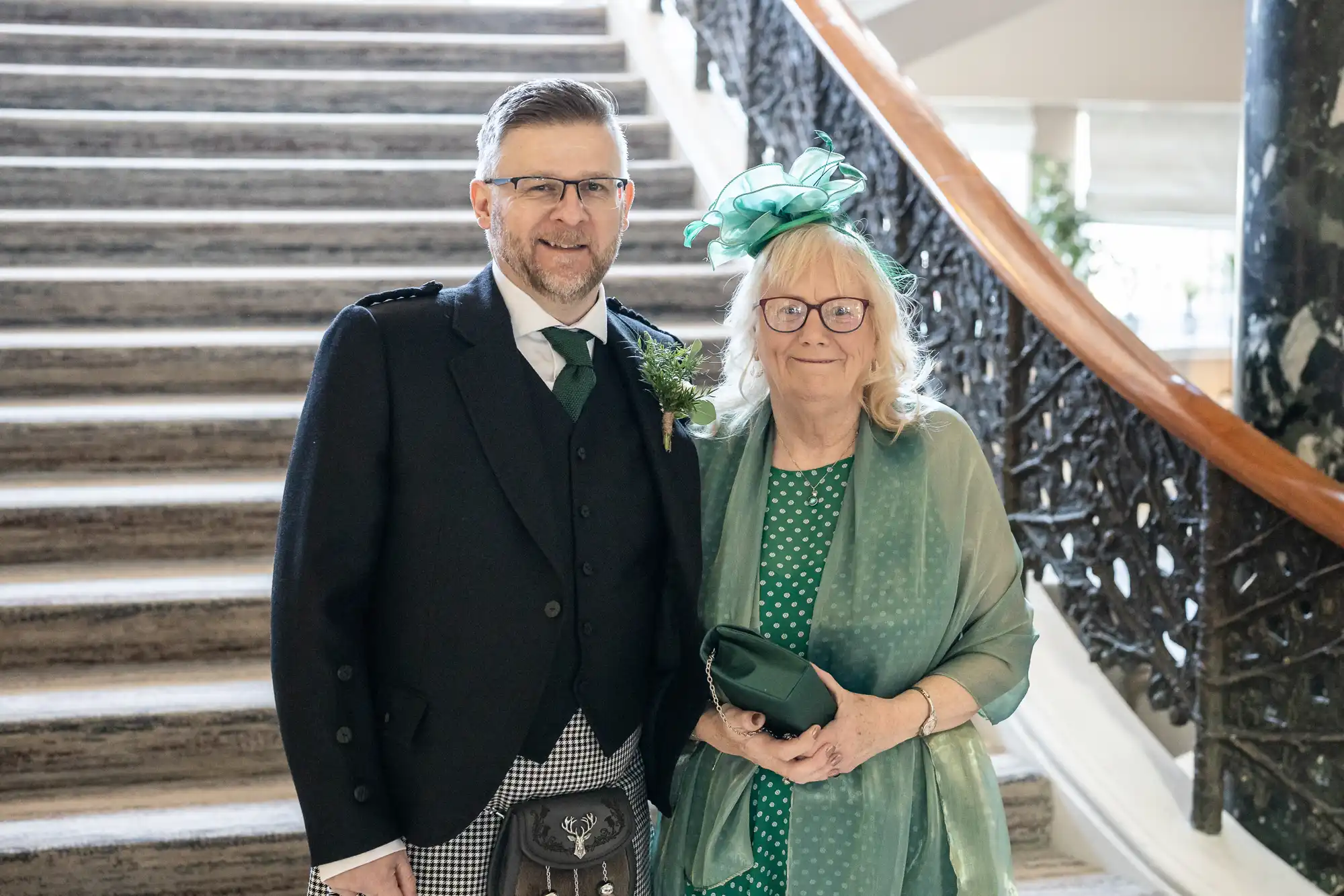 A man in a formal Scottish outfit and a woman in a green dress and hat stand together in front of a staircase.