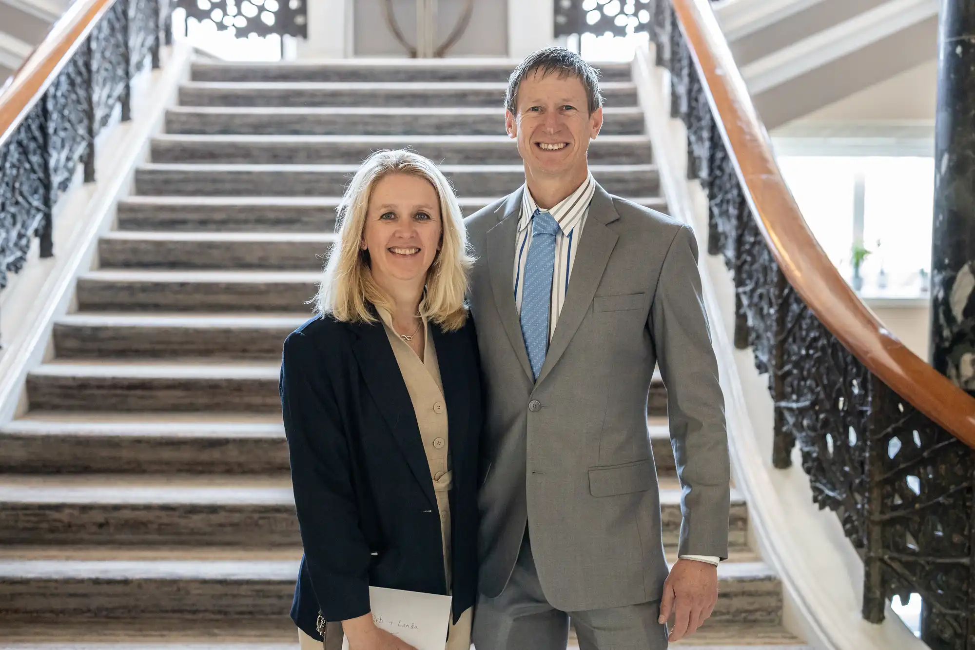 A woman and a man stand side by side, both smiling, in front of a staircase with ornate railings. The woman wears a suit jacket, and the man is dressed in a grey suit.