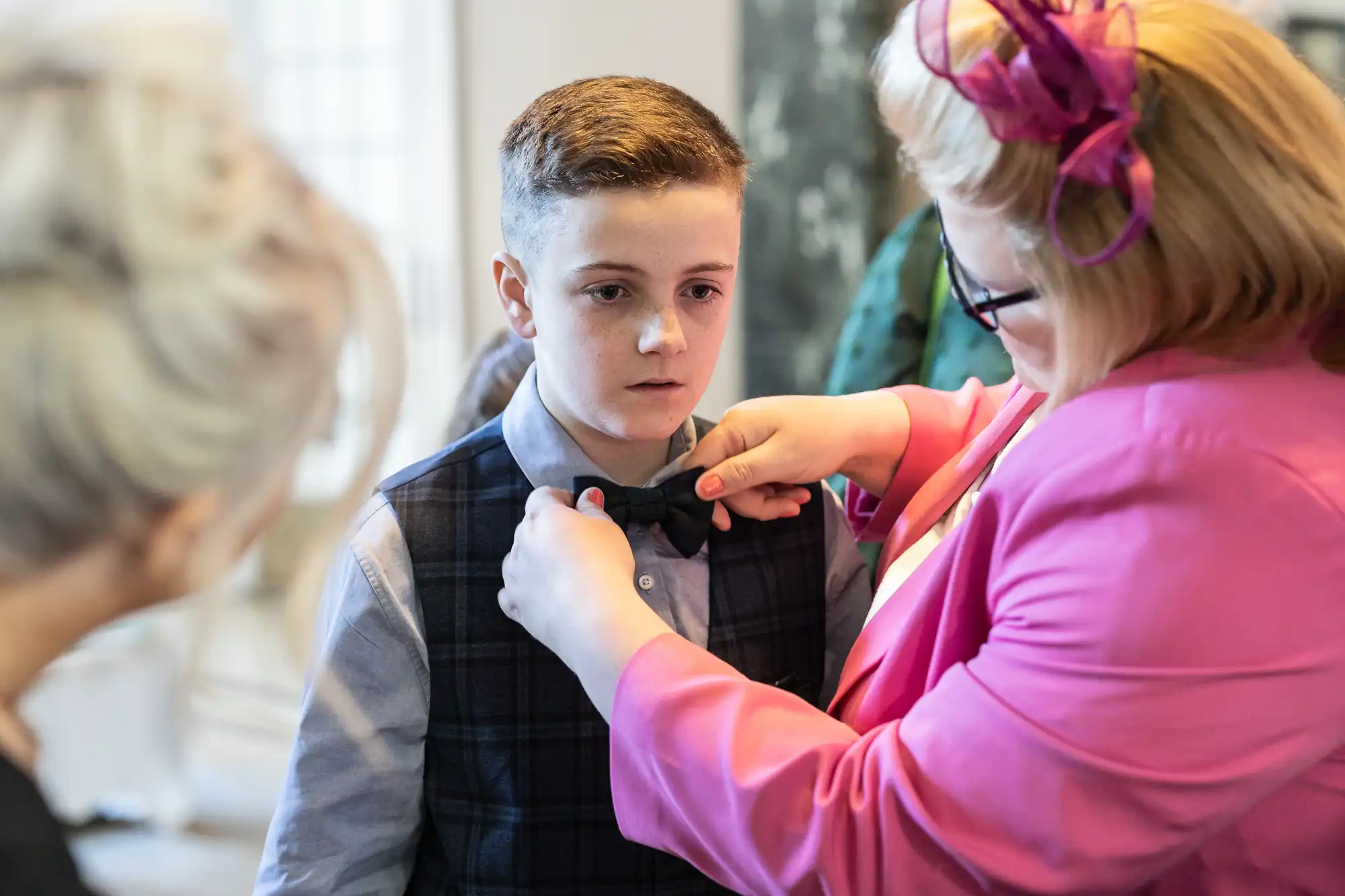 A boy in formal attire has his bow tie adjusted by a person in a pink jacket during an indoor event.